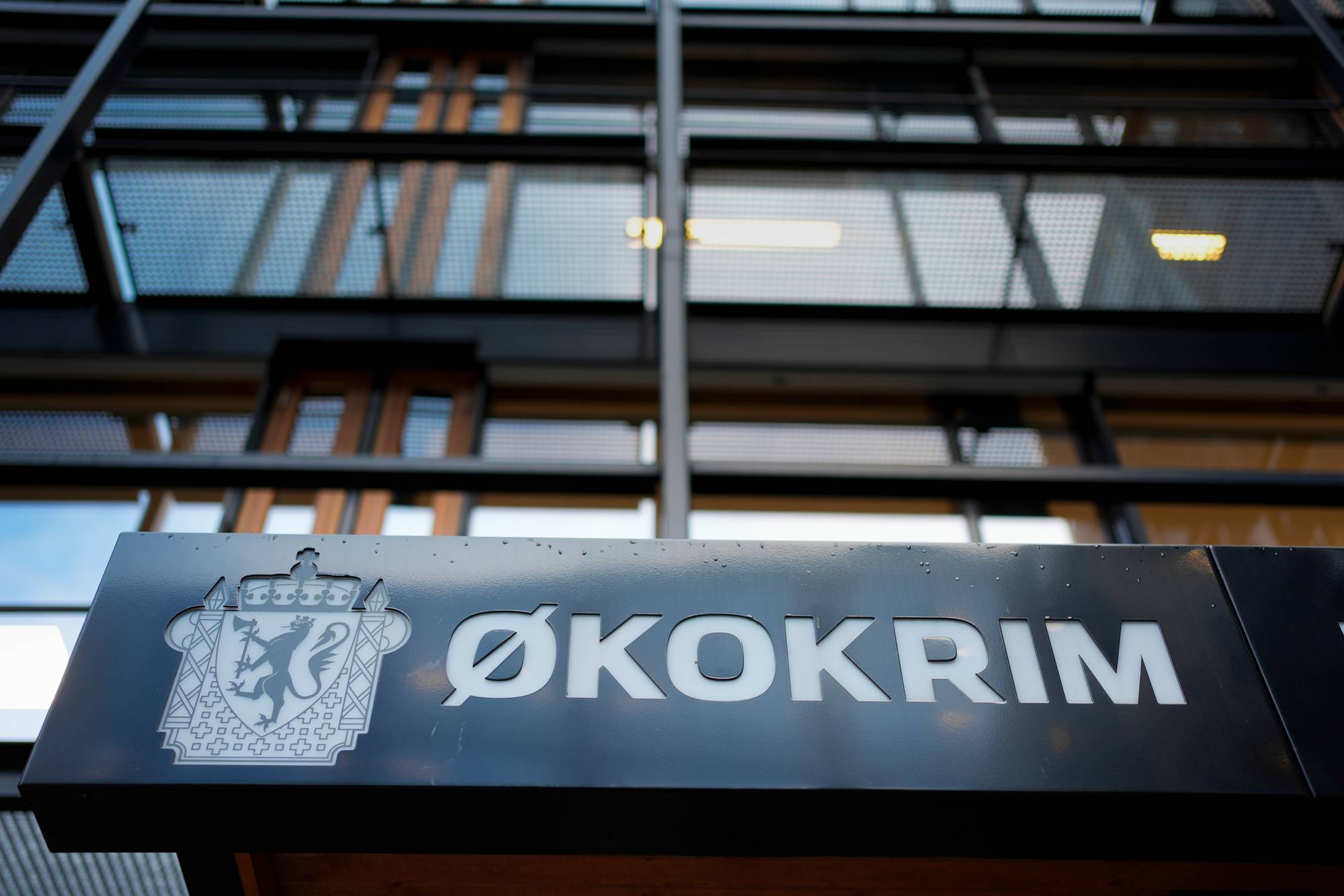 Bildet viser skiltet til Økokrim, med en detaljert logo og teksten "Økokrim" i store bokstaver. Skiltet henger på en moderne bygning med glassfasade og metallrammer. Det er synlig flere vinduer i bakgrunnen, som kan indikere et kontorlandskap. Været ser ut til å være klart, noe som bidrar til belysningen av området. (Bildebeskrivelsen er laget av en KI-tjeneste)