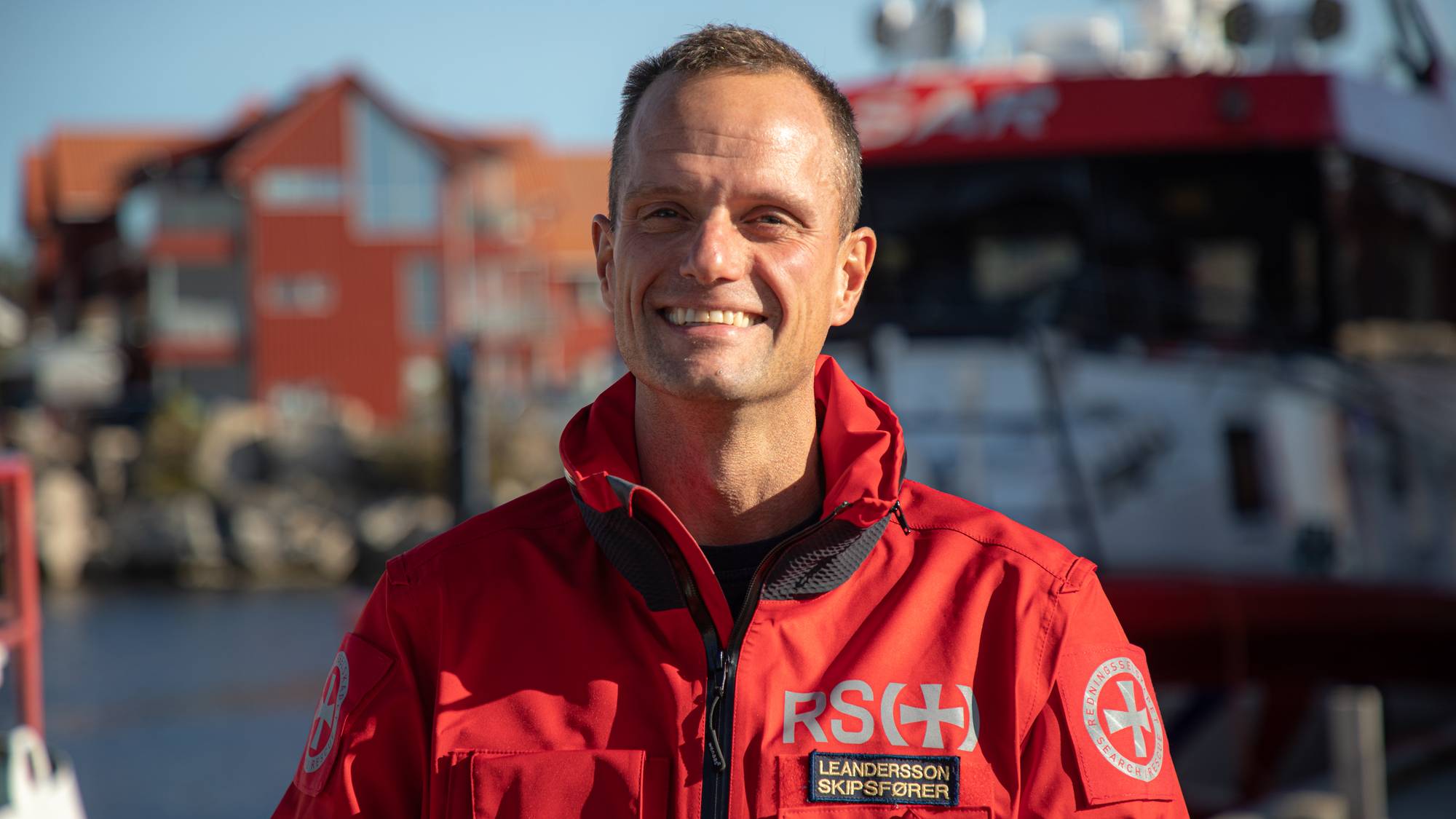 Blid mann i rød kjeledress smiler, man kan se redningsskøyta i bakgrunnen