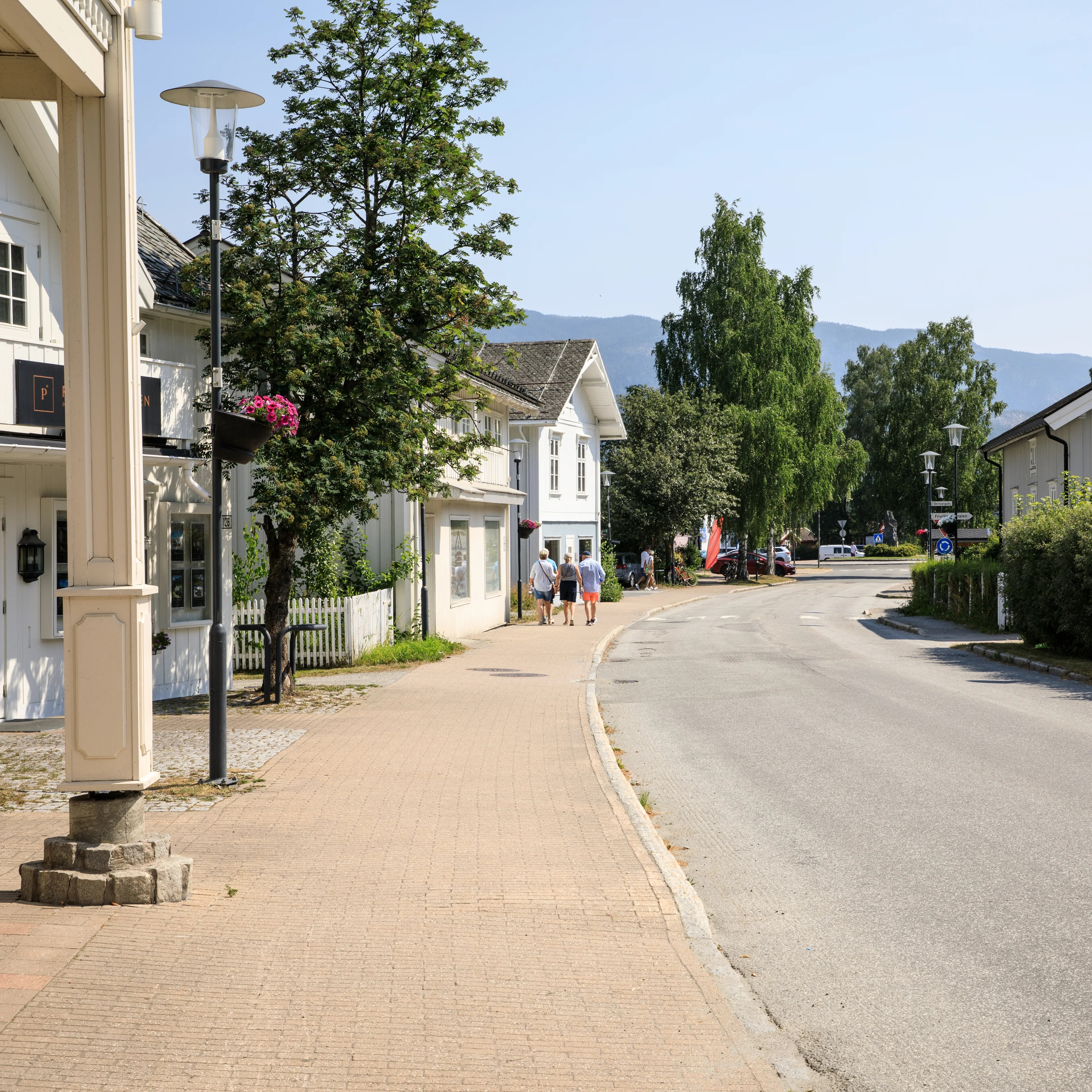 Tre personer går langs fortauet i en gate med hvit trehusbebyggelse på en solfylt dag. (Bildebeskrivelsen er laget av en KI-tjeneste)