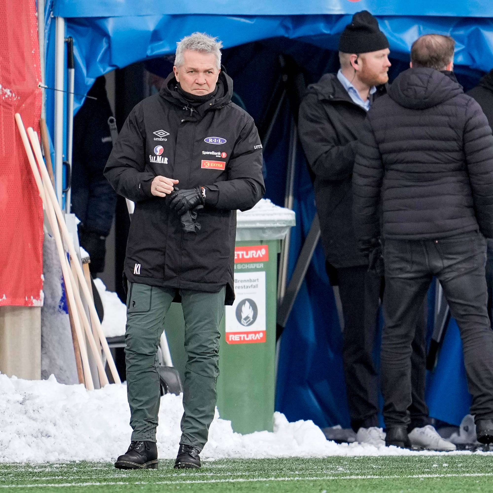 Flere personer står tett sammen ved en fotballbane. En mann i svart jakke og grønne bukser står lengst frem og ser mot høyre. Ved siden av ham står andre menn i yttertøy. Det ligger snø på bakken. (Bildebeskrivelsen er laget av en KI-tjeneste)