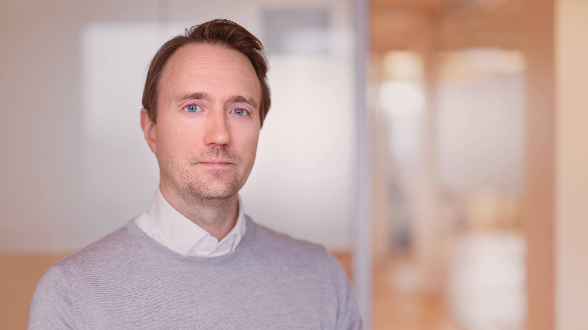 En mann med kort brunt hår og hvit skjorte under en grå genser ser alvorlig i kamera. I bakgrunn er lyse kontorlokaler.