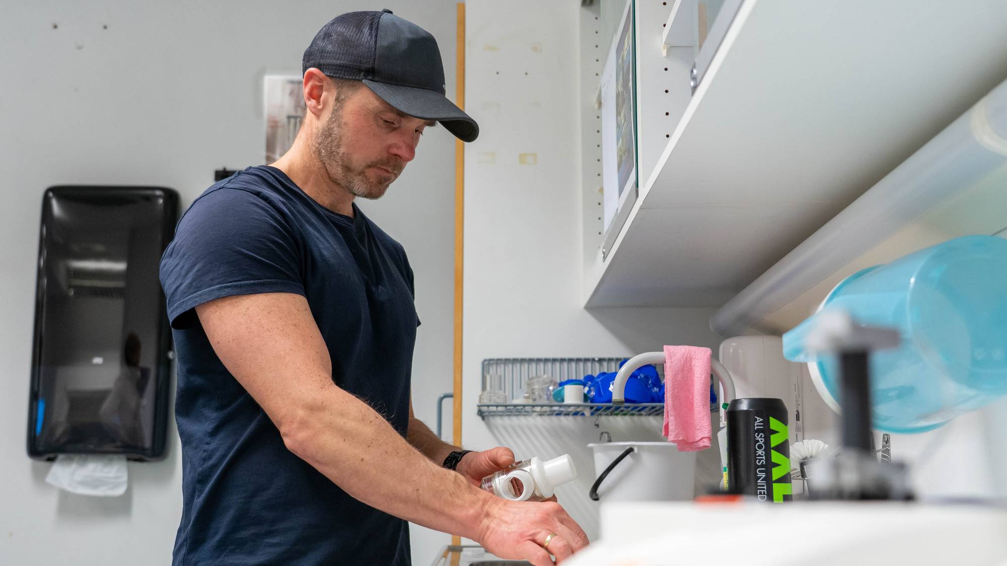 En mann med caps og mørk t-skjorte jobber over en vask. Han holder en hvit plastkomponent i hendene. Bak ham er det hyller med diverse gjenstander. Et svart dispensere for papirhåndklær henger på veggen til venstre.