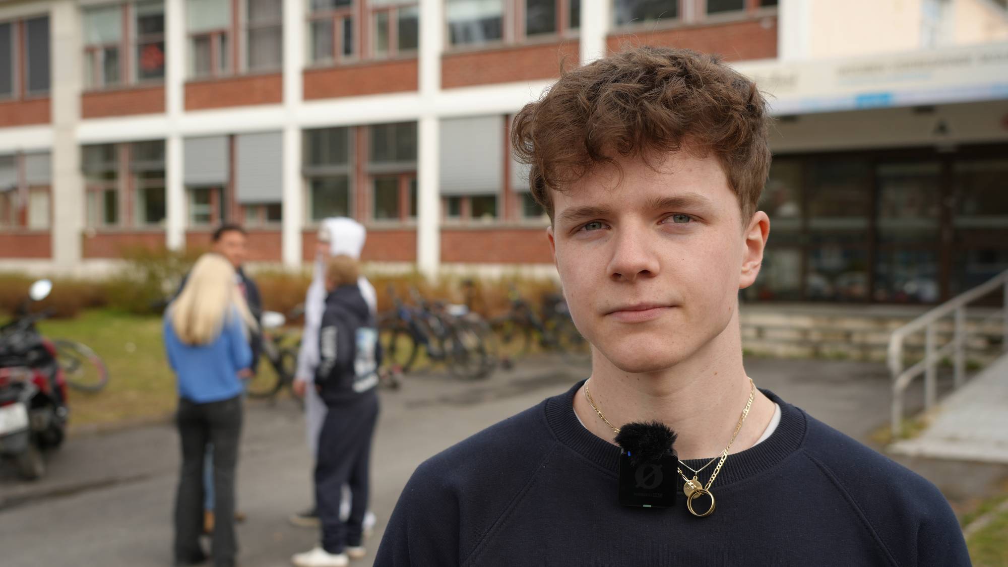 En mann som står forran et skolebygg med flere folk i bakgrunnen. 