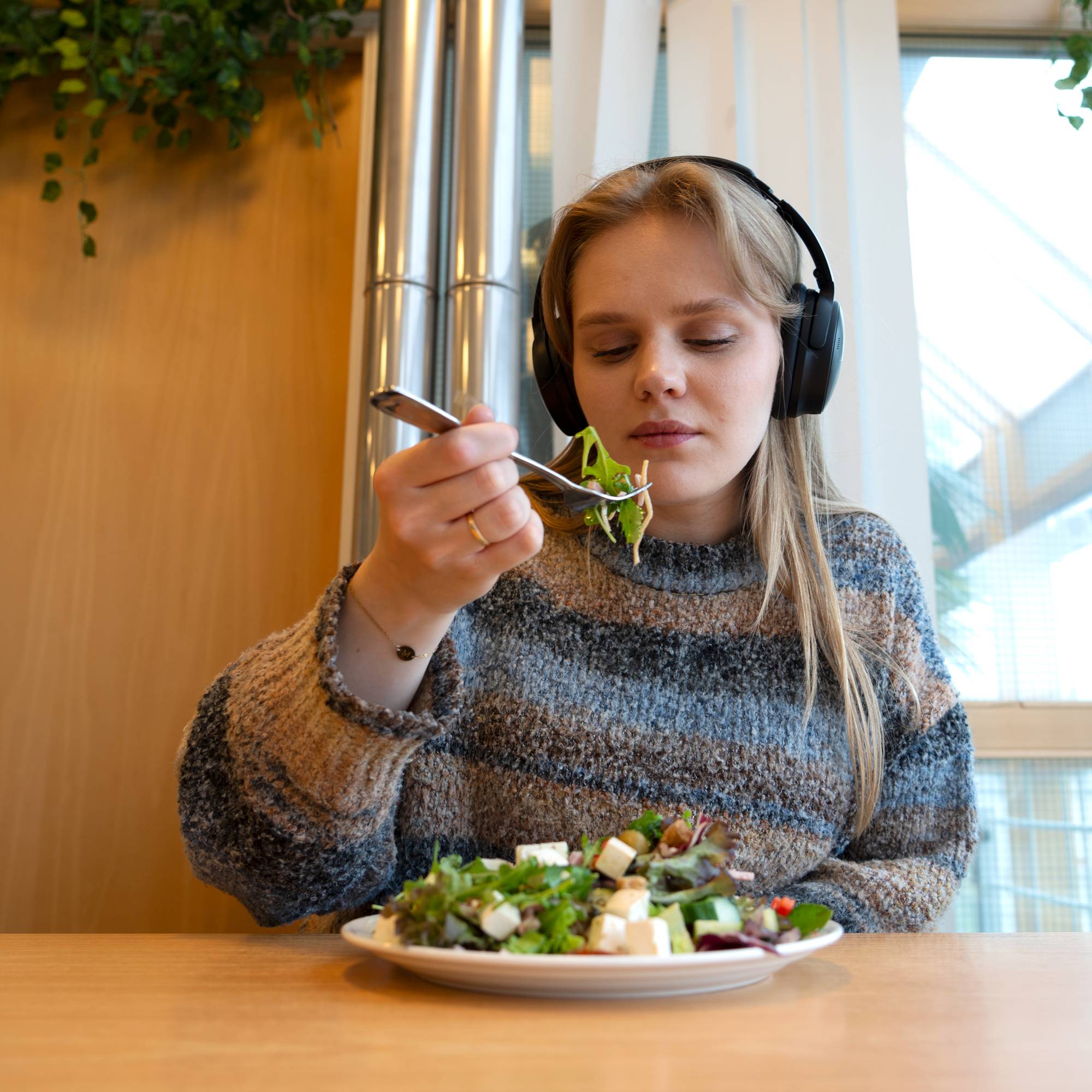 Kvinne spiser og hører musikk med hodetelefoner. Hun sitter ved et bord.