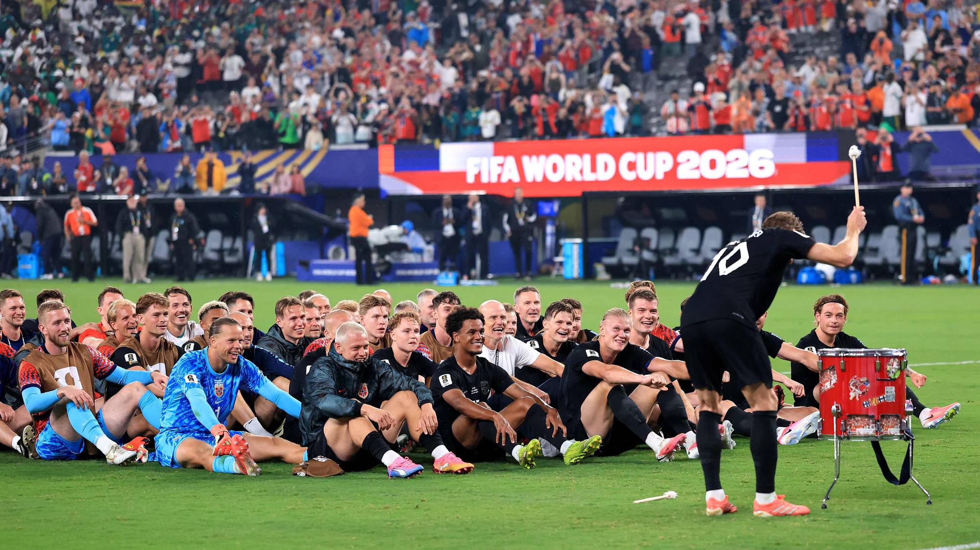 Et fotballag feirer på et jorde, med publikum i bakgrunnen. En mann slår på en tromme, mens andre sitter på bakken. En reklame for FIFA WORLD CUP 2026 ses i bakgrunnen. (Bildebeskrivelsen er laget av en KI-tjeneste)