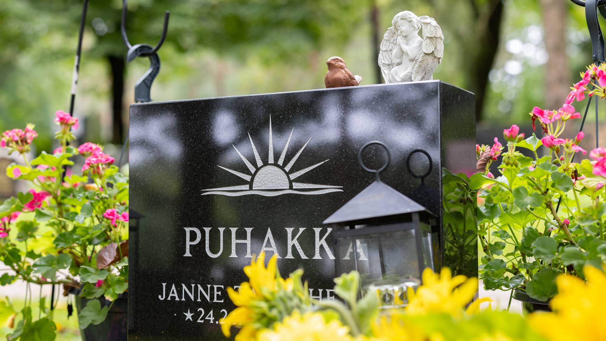 En gravstøtte på en kirkegård. Det er friske blomster ved graven i ulike farger. På steinen kan vi lese deler av navnet til han som er begravet, Puhakka.