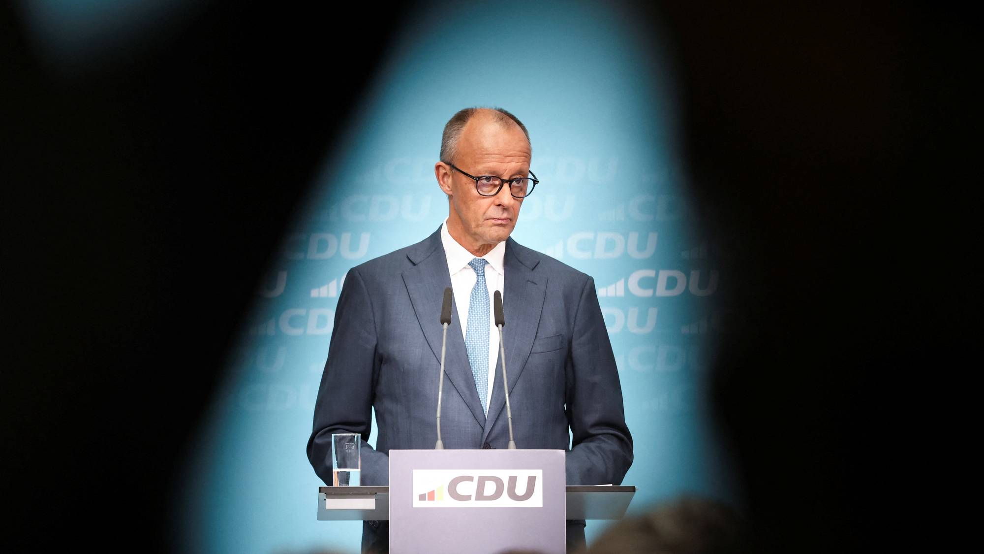 Friedrich Merz står bak en talerstol med CDU-logo foran en lyseblå bakgrunn. (Bildebeskrivelsen er laget av en KI-tjeneste)