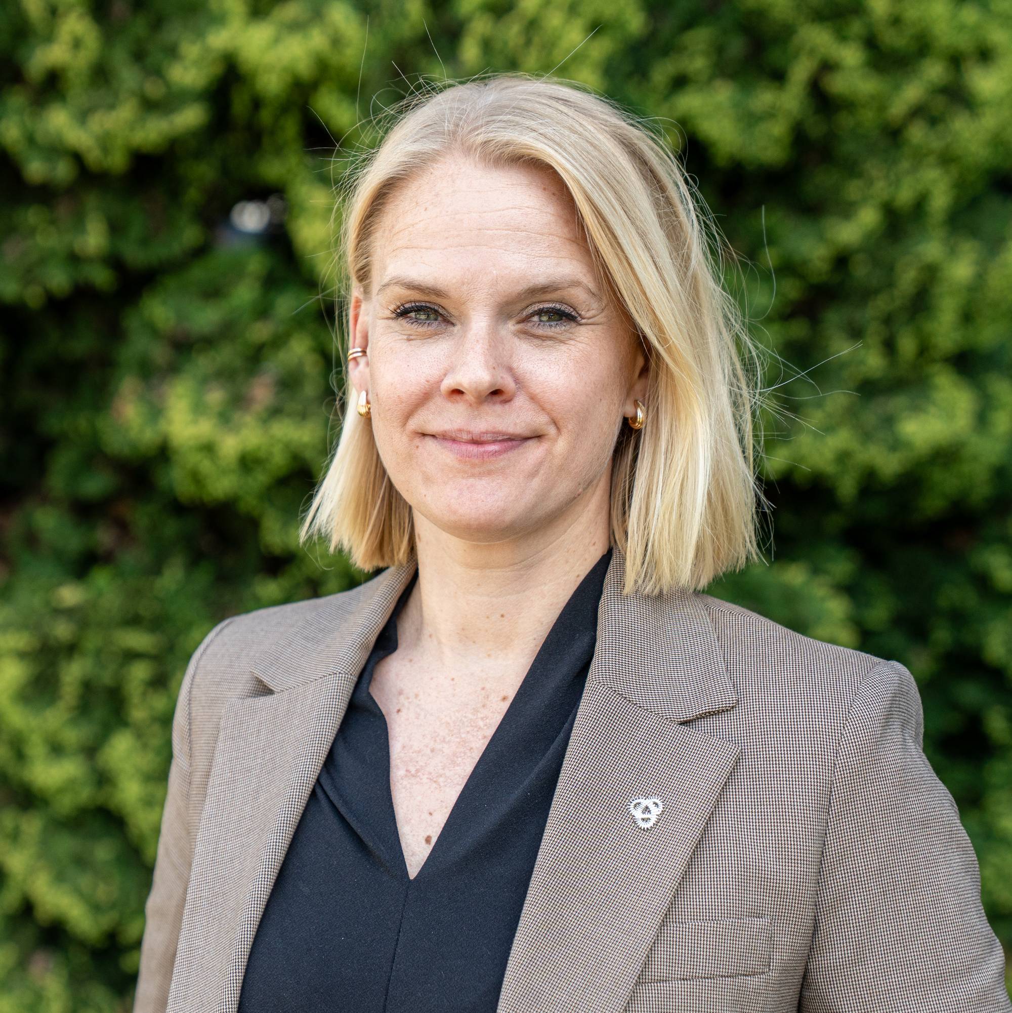 En kvinne i jakke ser mot kameraet. Hun står foran en grønn busk. Kvinnen har blondt hår og smiler.