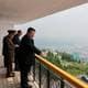 Reuters Flere menn står på en balkong med utsikt over et grønt landskap. Den ene mannen peker ut over utsikten, mens en annen skriver ned notater. Omgivelsene inkluderer trær og bygninger i det fjerne, samt en vei som strekker seg langs landskapet. Balkongen er innrammet med et gelender og har gulv av fliser.