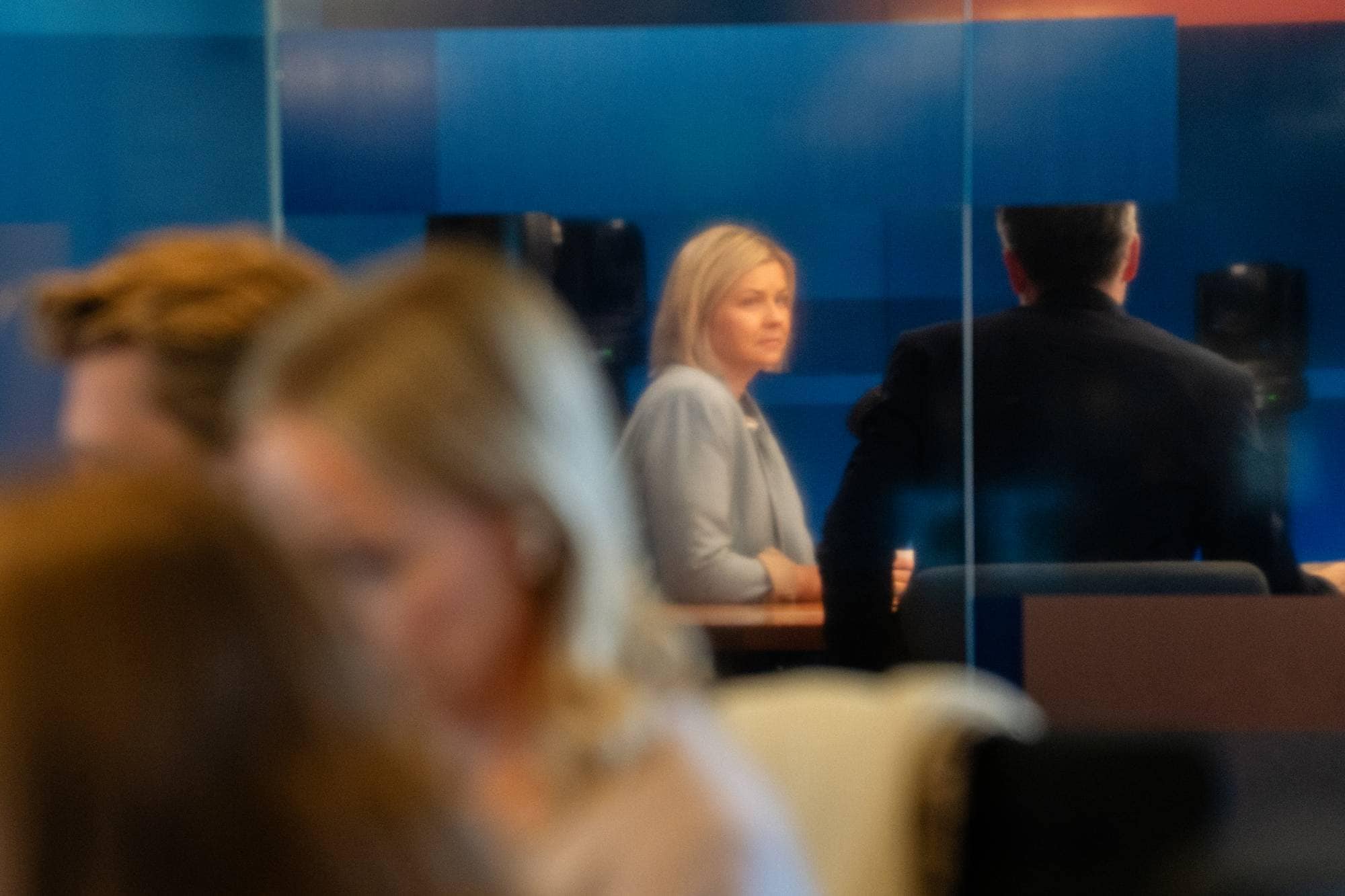 En kvinne og en mann sitter ved et bord, med flere personer i forgrunnen. Kvinnen har blondt hår og har på seg en lyseblå jakke. Mannen sitter med ryggen til kameraet, kledd i en mørk dressjakke. Bak dem er en blå vegg med noen firkantede felt. (Bildebeskrivelsen er laget av en KI-tjeneste)
