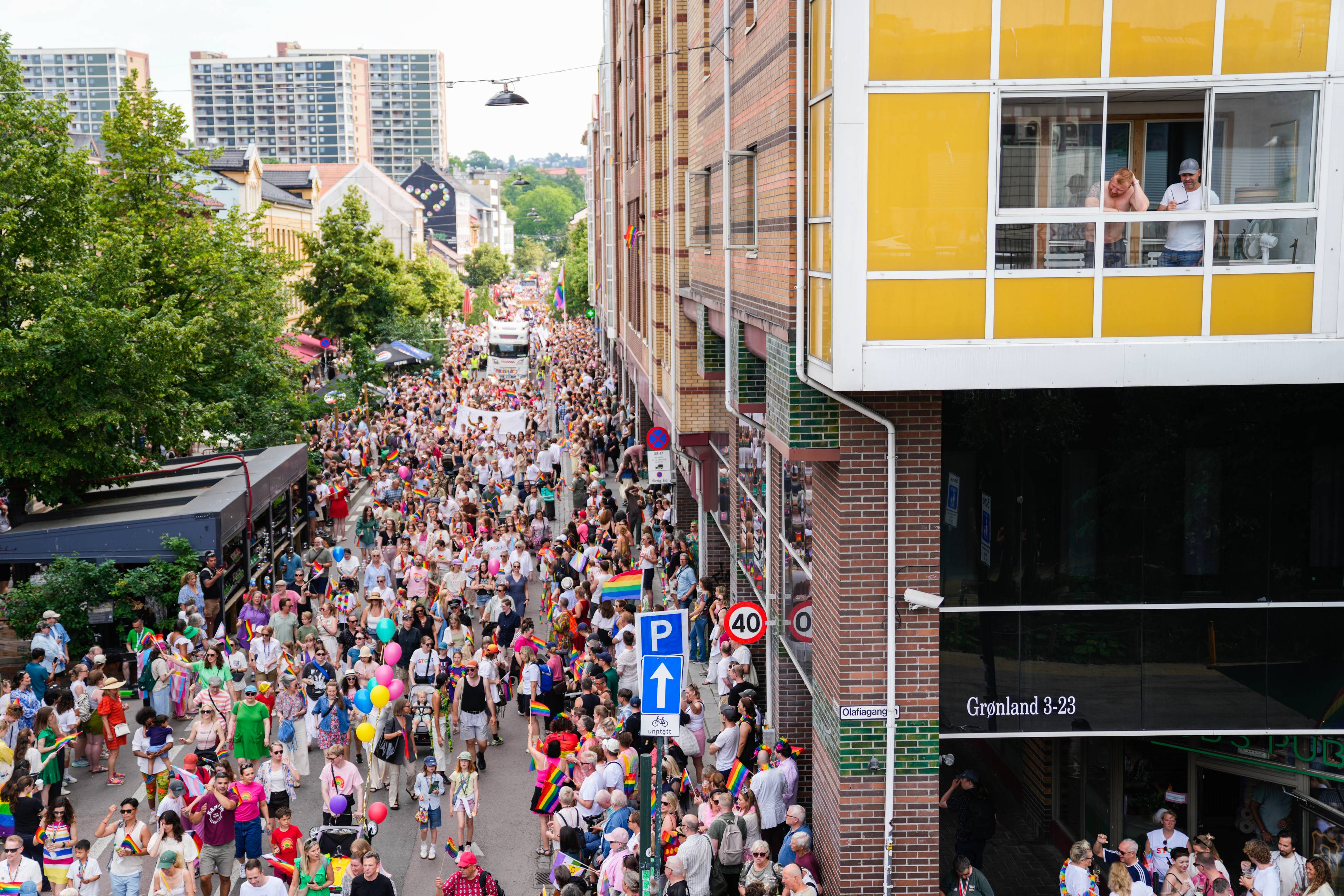 En stor folkemengde går i Pride-parade gjennom en bygate med bygninger på sidene. (Bildebeskrivelsen er laget av en KI-tjeneste)