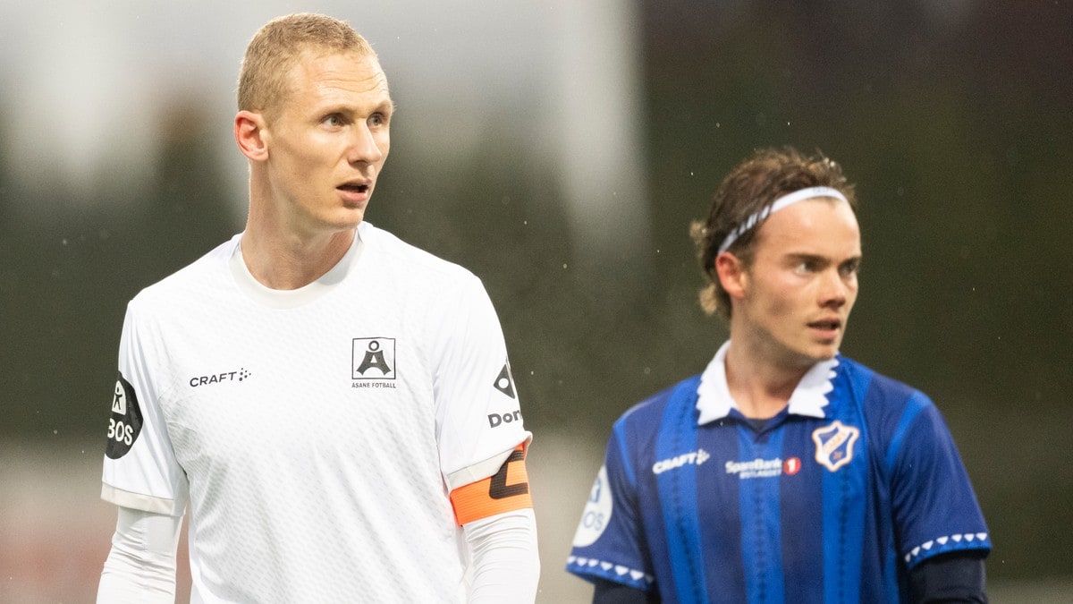 To fotballspillere i drakter fra Åsane og et annet lag står på banen under en kamp. (Bildebeskrivelsen er laget av en KI-tjeneste) FOTO: Annika Byrde/NTB