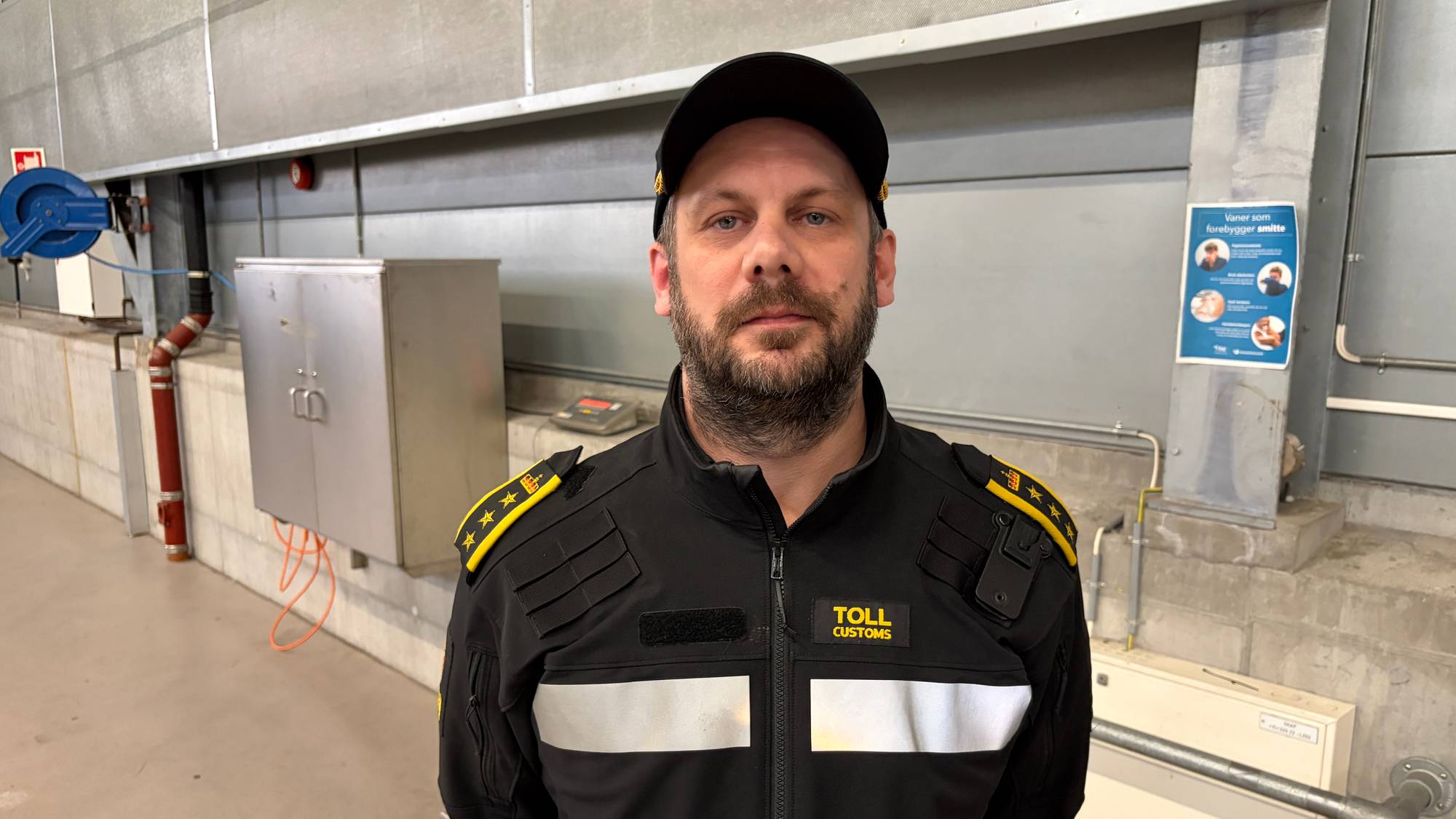 Tollbetjent i uniform står foran en betongvegg i det som ser ut som et tollager eller godsterminal. (Bildebeskrivelsen er laget av en KI-tjeneste)