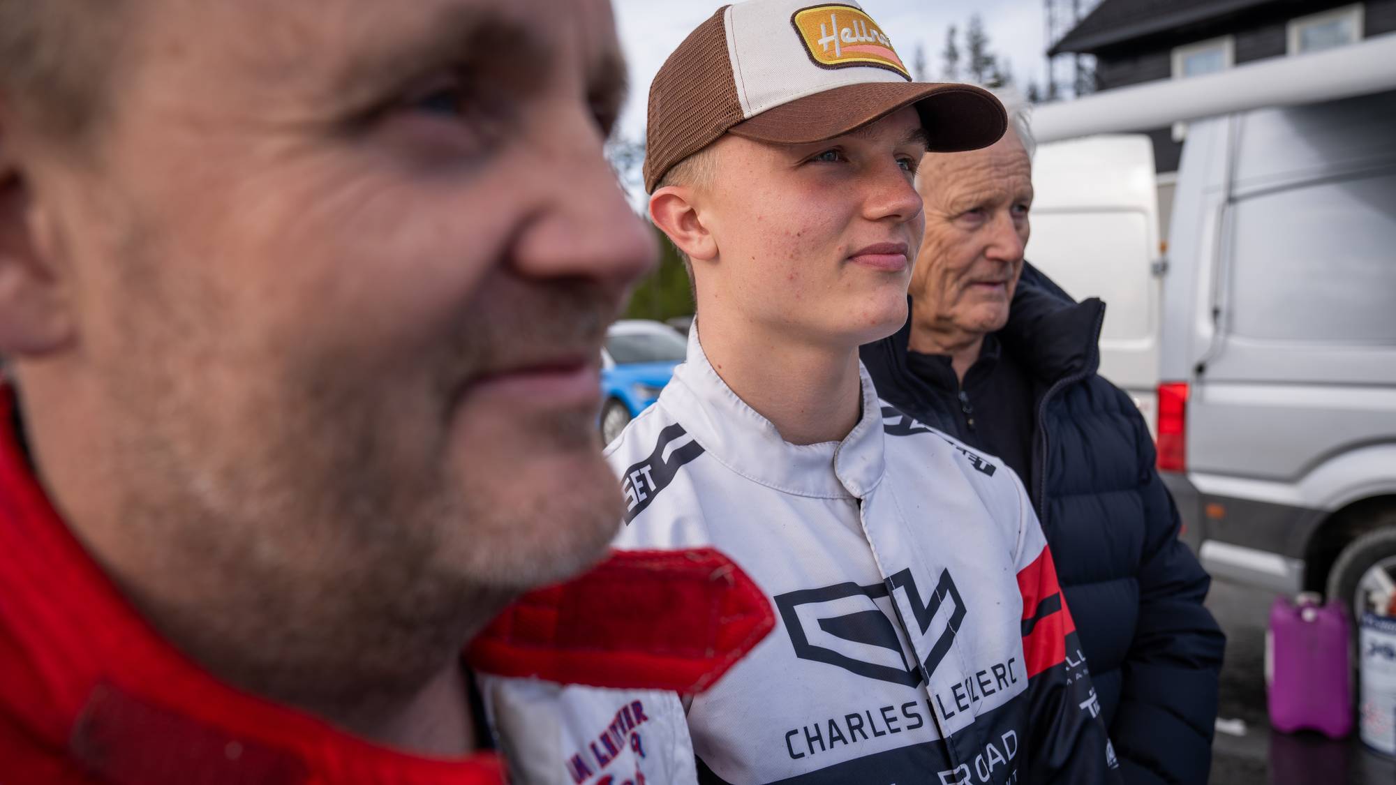 Tre menn i racerdrakter står side om side og ser mot siden. En ung mann i midten har på seg en caps med teksten "Hello". Bak dem står hvite og sølvfargede varebiler. (Bildebeskrivelsen er laget av en KI-tjeneste)