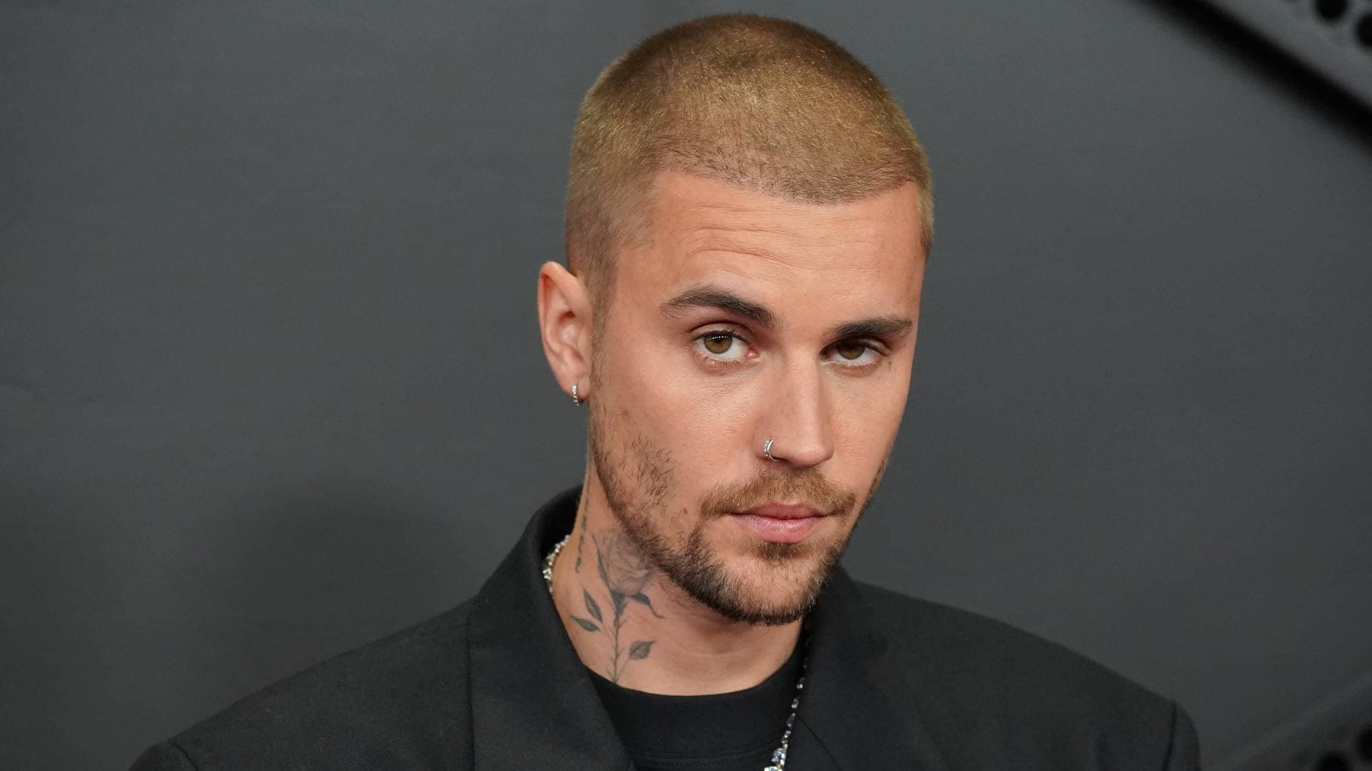 Portrett av artisten Justin Bieber. Han har kortklipt blondt hår og er ikledd en svart blazer. Bieber har flere tatoveringer, blant annet på halsen. Bakgrunnen er mørkegrå. (Bildebeskrivelsen er laget av en KI-tjeneste)