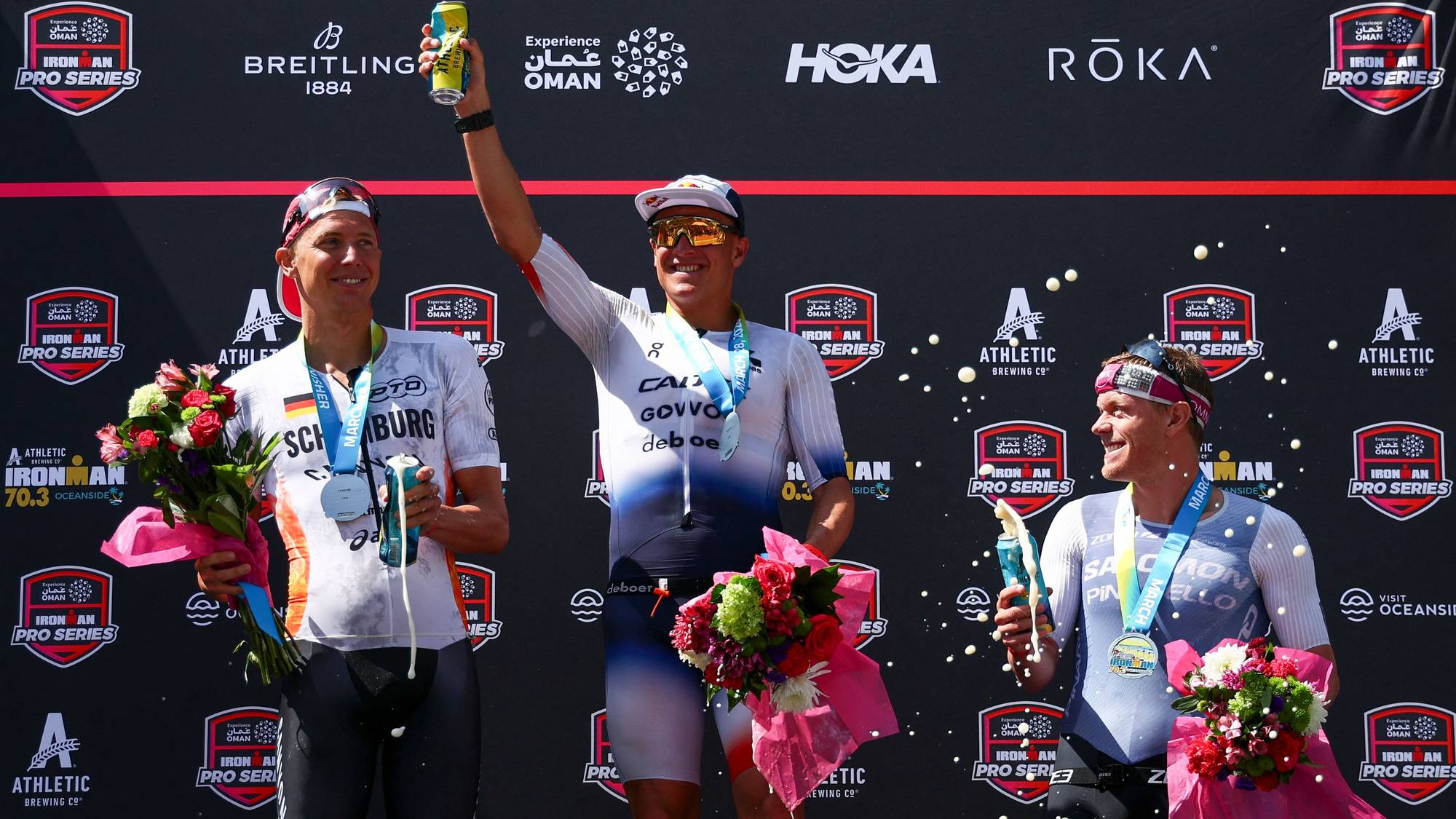 Tre menn på en seierspodium holder blomsterbuketter og drikke. De har medaljer rundt halsen. En av mennene sprayer champagne. Bak dem er en svart bakgrunn med sponsorlogoer. (Bildebeskrivelsen er laget av en KI-tjeneste)