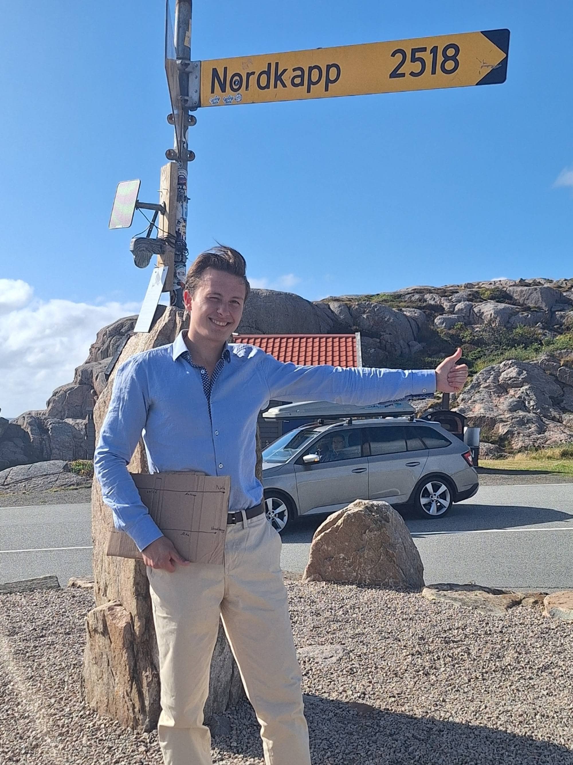 Alexander Sirris står framfor et skilt i Lindesnes som viser at Nordkapp er 2518km unna. Han holder haiketommelen opp og smiler.