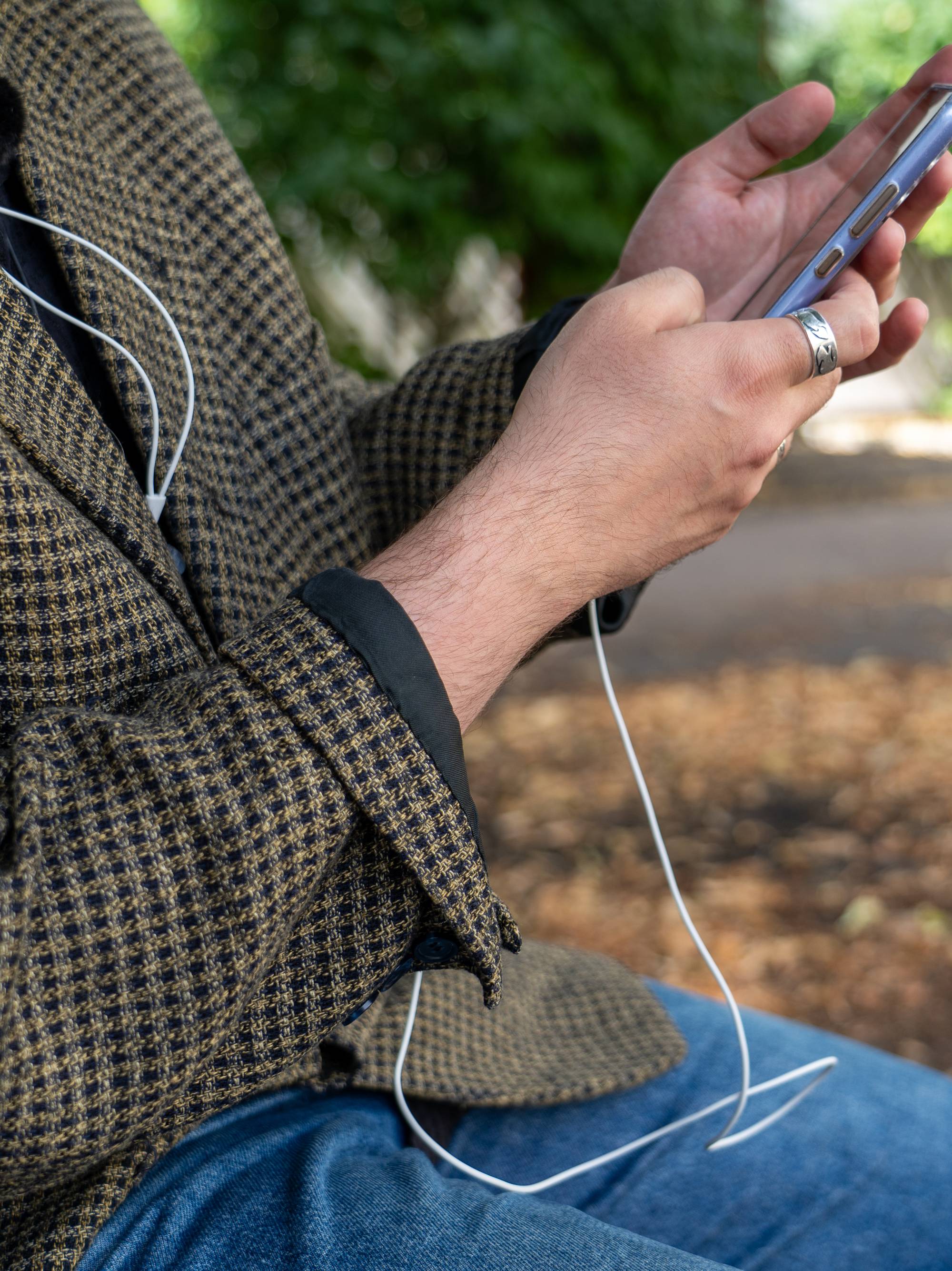 Person i rutete jakke sitter utendørs og bruker en smarttelefon med ørepropper. (Bildebeskrivelsen er laget av en KI-tjeneste)