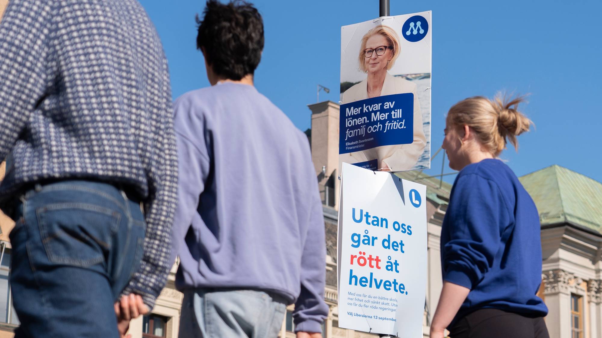 Folk går forbi valgplakater for Moderaterna og Liberalerna på en bygate i solskinn. (Bildebeskrivelsen er laget av en KI-tjeneste)