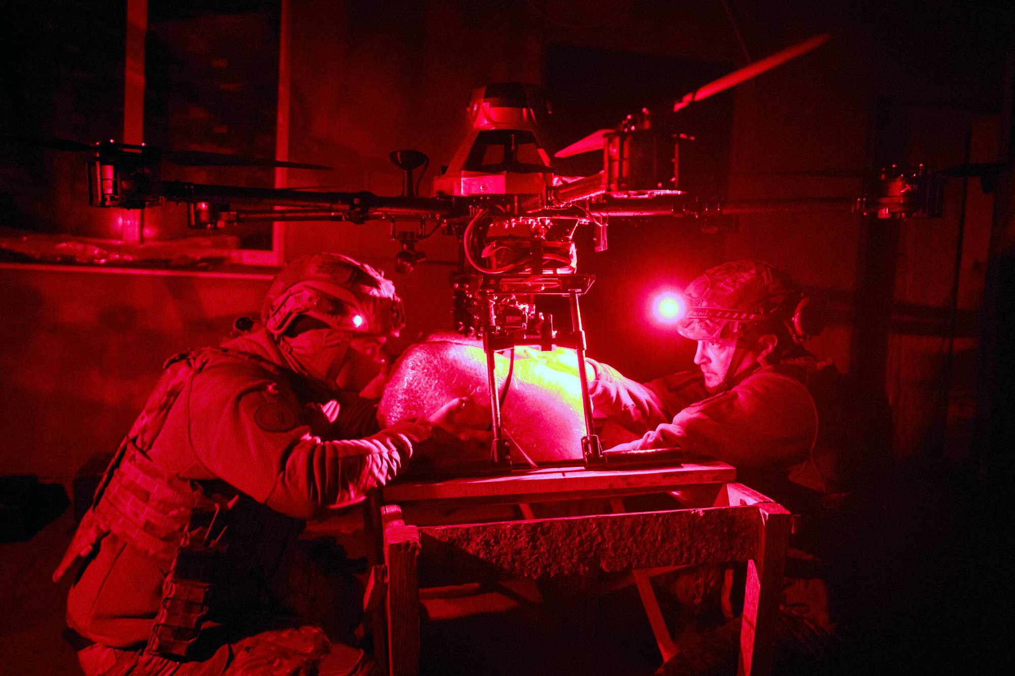To soldater jobber med en drone i et rom med rødt lys. Den ene soldaten kneler foran et bord. Begge er iført hjelm og vest. En stor drone med propeller er plassert over bordet. (Bildebeskrivelsen er laget av en KI-tjeneste)