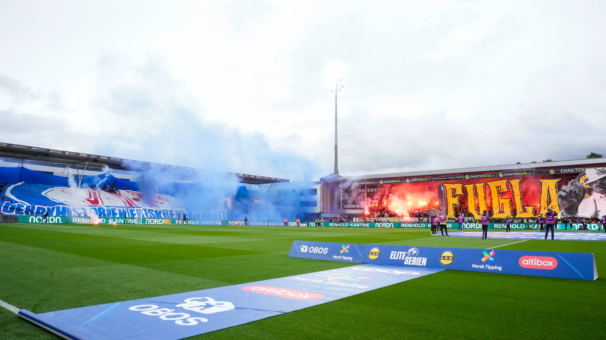 Supportere lager bluss- og røykeffekter på hver sin tribune under en fotballkamp på en stadion. (Bildebeskrivelsen er laget av en KI-tjeneste)