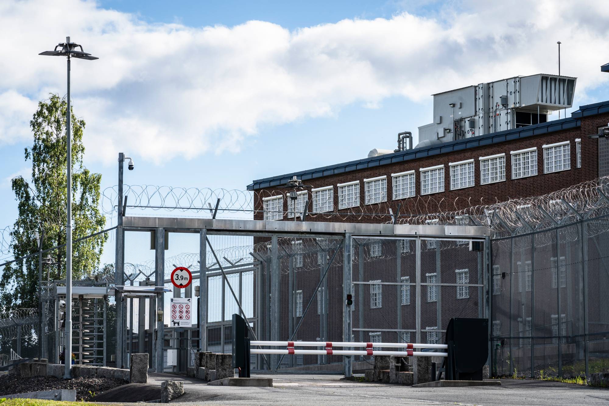 Fengselsport med piggtrådgjerder og en teglsteinsbygning i bakgrunnen. (Bildebeskrivelsen er laget av en KI-tjeneste)