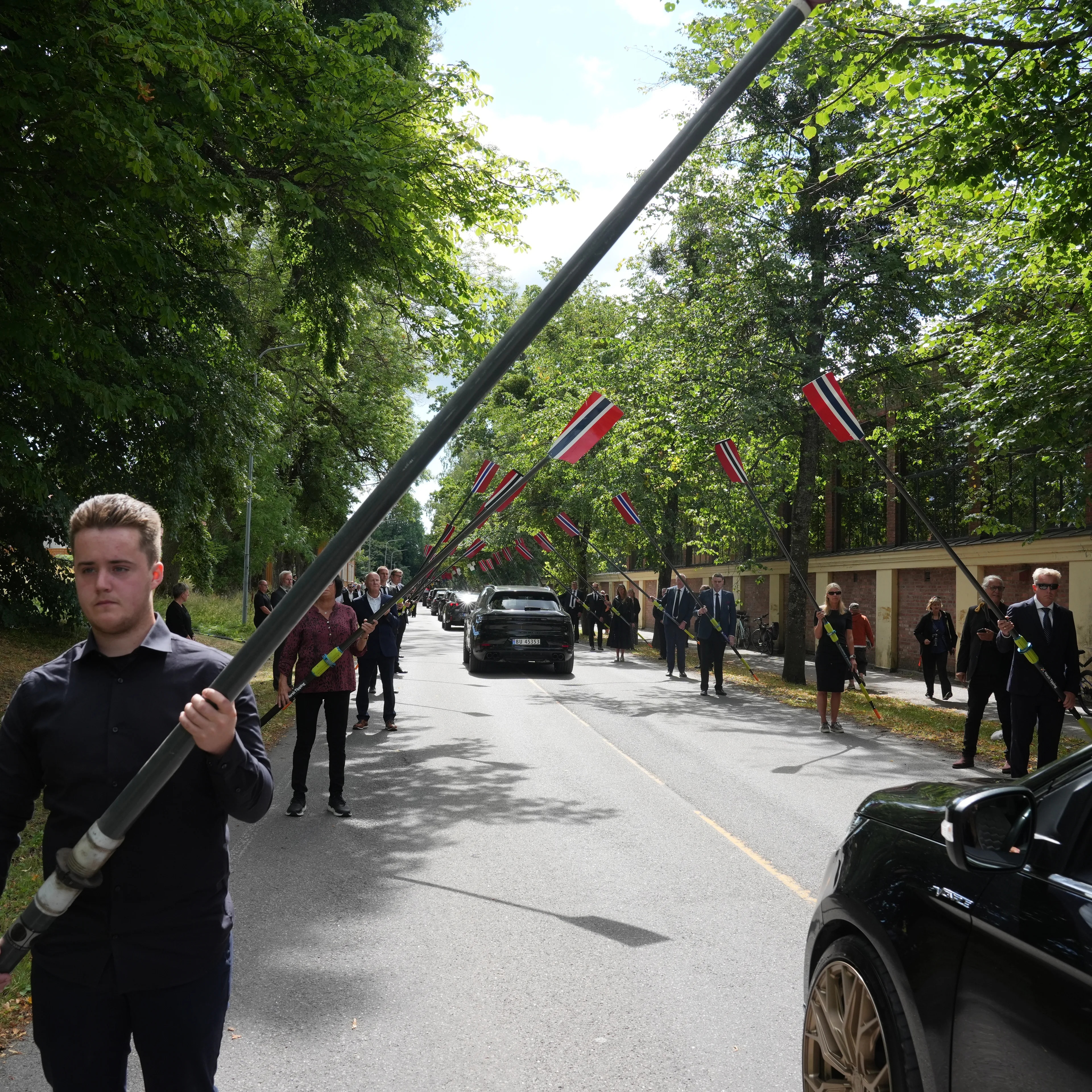 Personer står langs veien og holder oppe årer med norske flagg under en seremoni.
