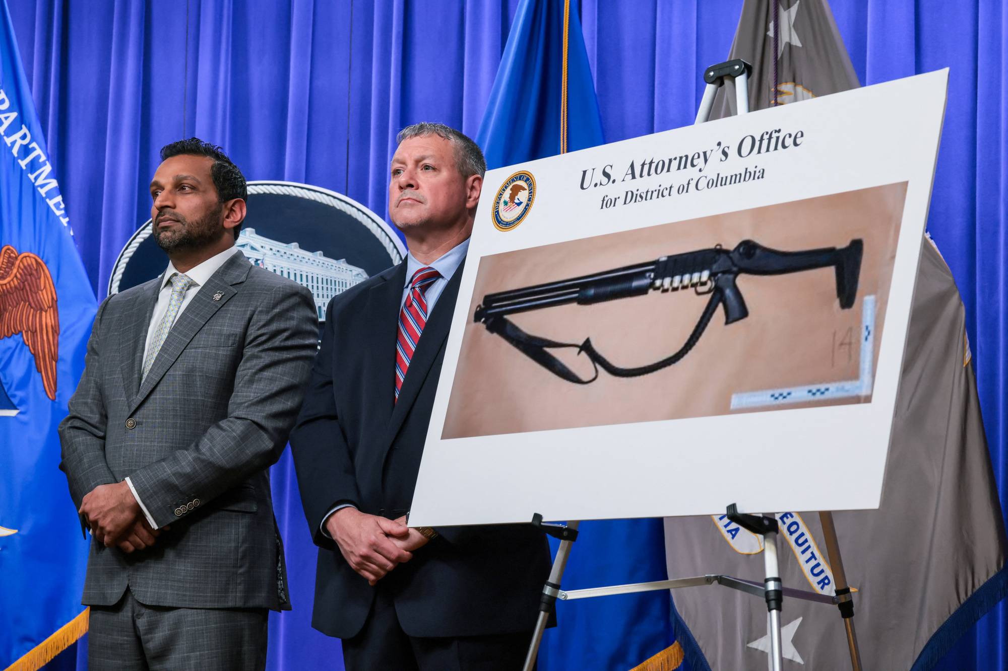 To menn står foran et blått forheng mens en tredje person holder opp et bilde av et våpen. Bildet viser et trykk av et våpen og teksten U.S. Attorney's Office for District of Columbia. Det er et amerikansk flagg på høyre side. (Bildebeskrivelsen er laget av en KI-tjeneste)