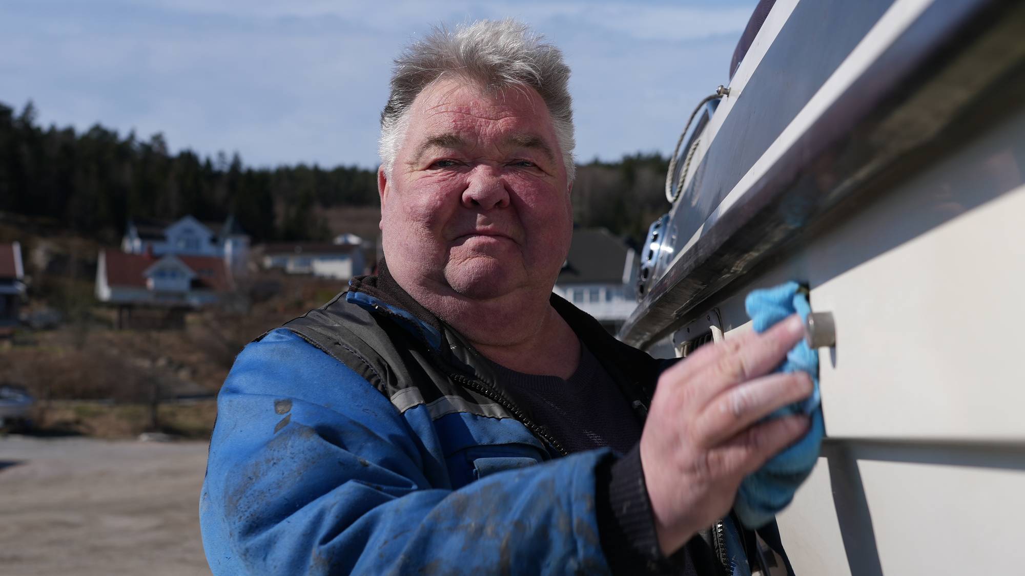 En mann med grått hår og blå jakke vasker en båt med en blå klut. Mannen ser inn i kamera. Bakgrunnen viser hus og trær. (Bildebeskrivelsen er laget av en KI-tjeneste)