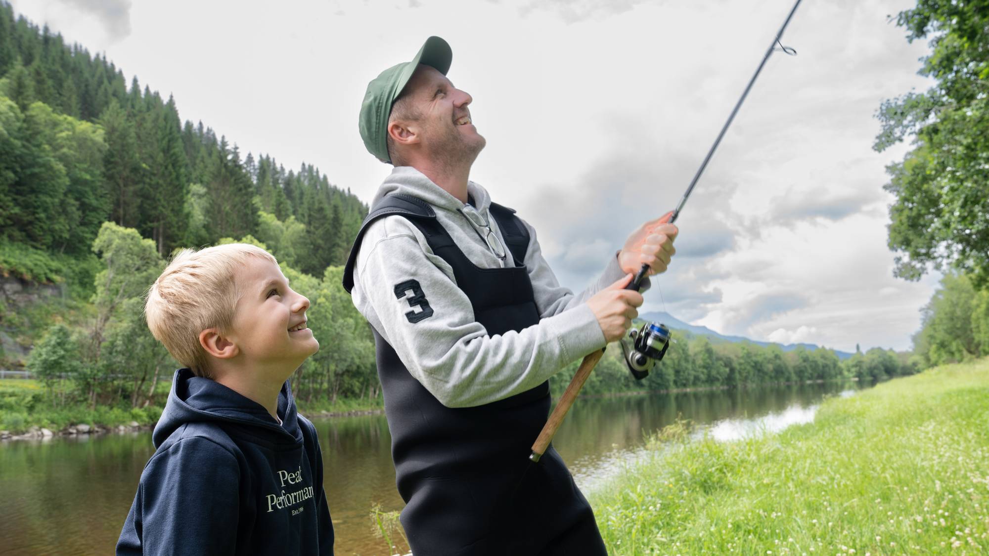 En mann og en gutt fisker sammen ved en elv omgitt av skog og grøntarealer. (Bildebeskrivelsen er laget av en KI-tjeneste)