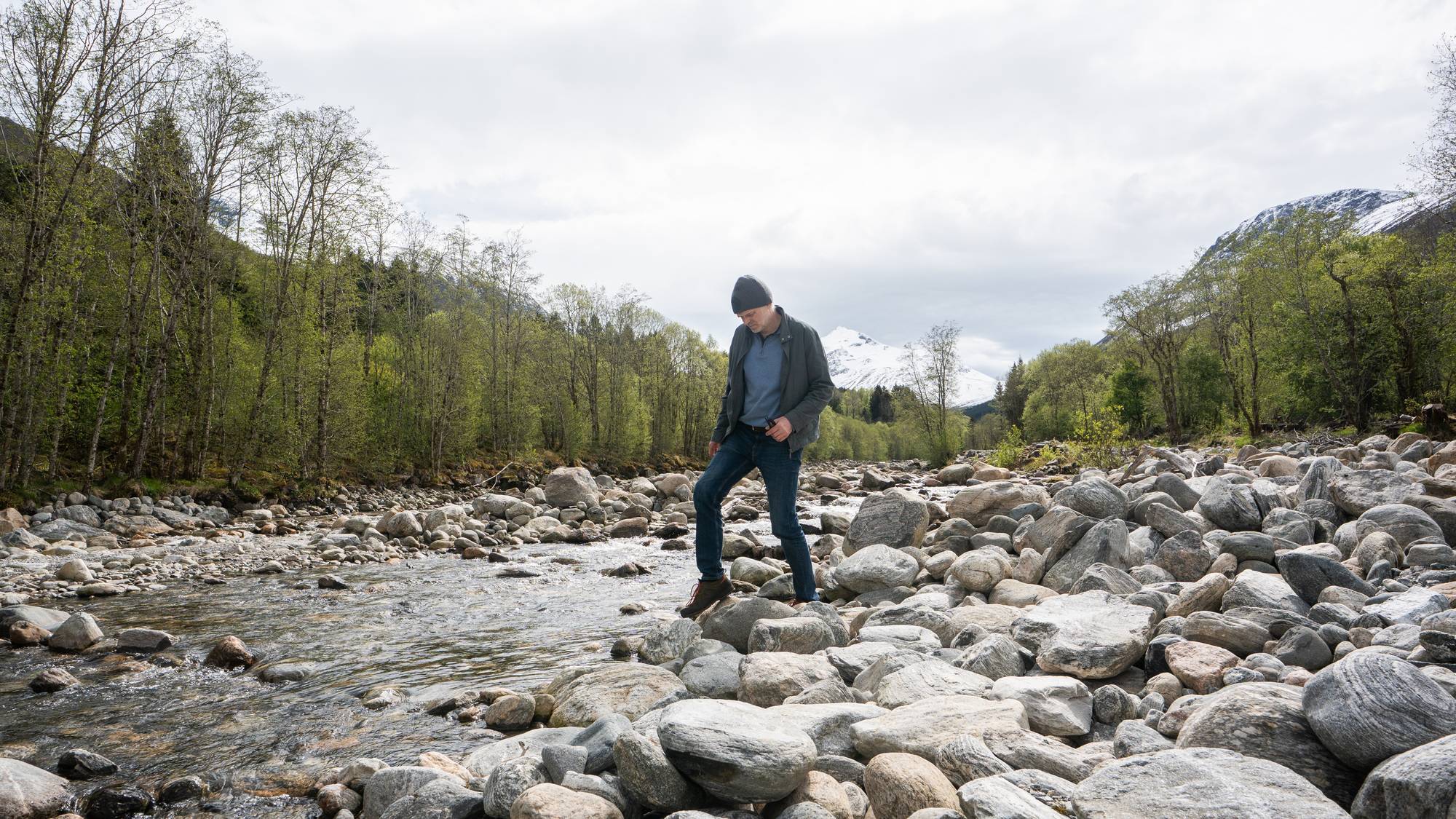 En mann står på steiner ved en elv. Mannen har på seg jakke, genser og jeans, og ser ned. Trær dekker sidene bak elven, og snødekte fjelltopper er synlige i bakgrunnen. (Bildebeskrivelsen er laget av en KI-tjeneste)