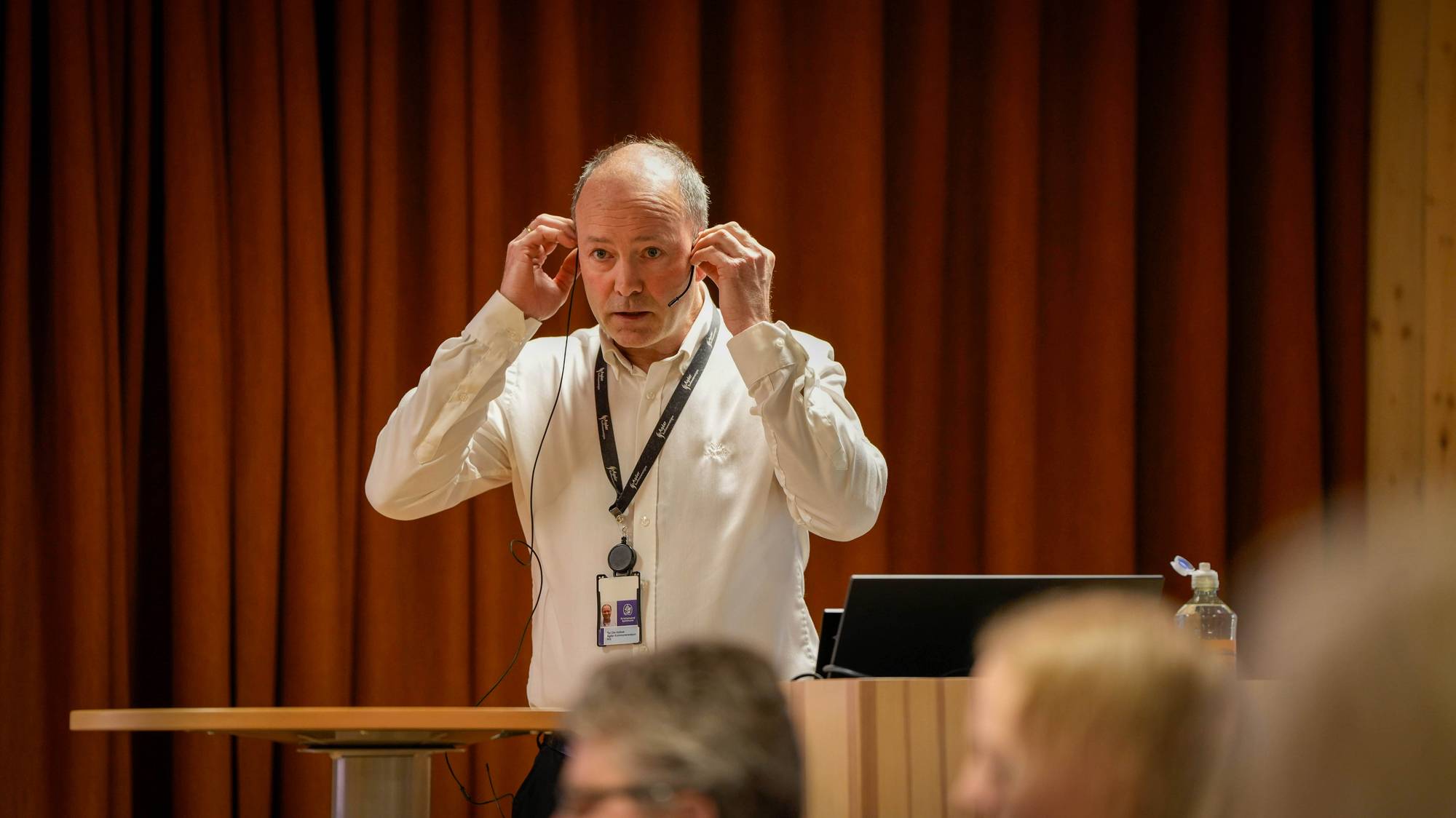 En mann i hvit skjorte justerer et headset. Han står ved et podium foran en brun bakgrunn. Til høyre for mannen er en dataskjerm og en flaske hånddesinfeksjon. Flere personer er synlige i forgrunnen. (Bildebeskrivelsen er laget av en KI-tjeneste)
