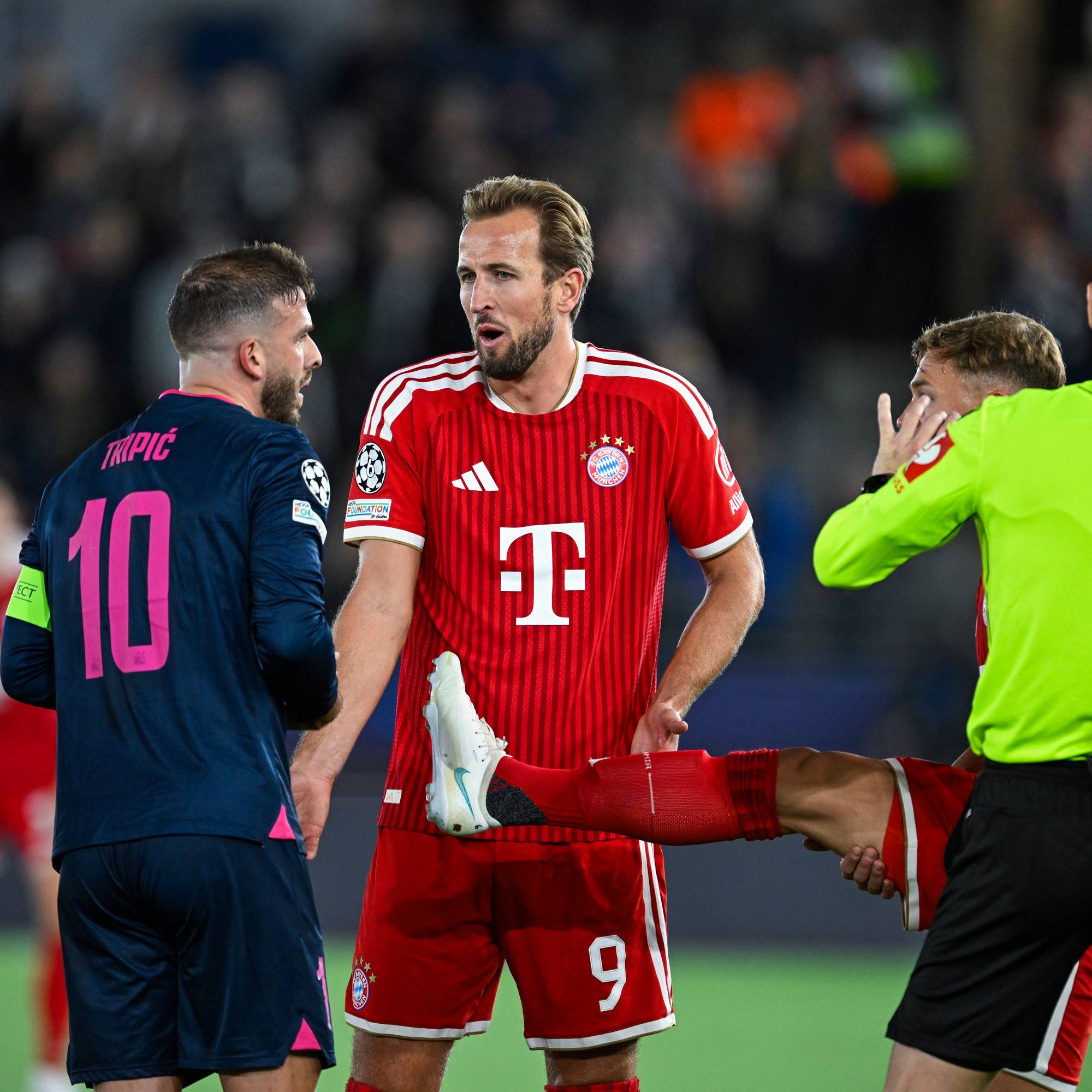 Harry Kane i Bayern München-drakt diskuterer med motspillere og dommer på en fotballbane. (Bildebeskrivelsen er laget av en KI-tjeneste)