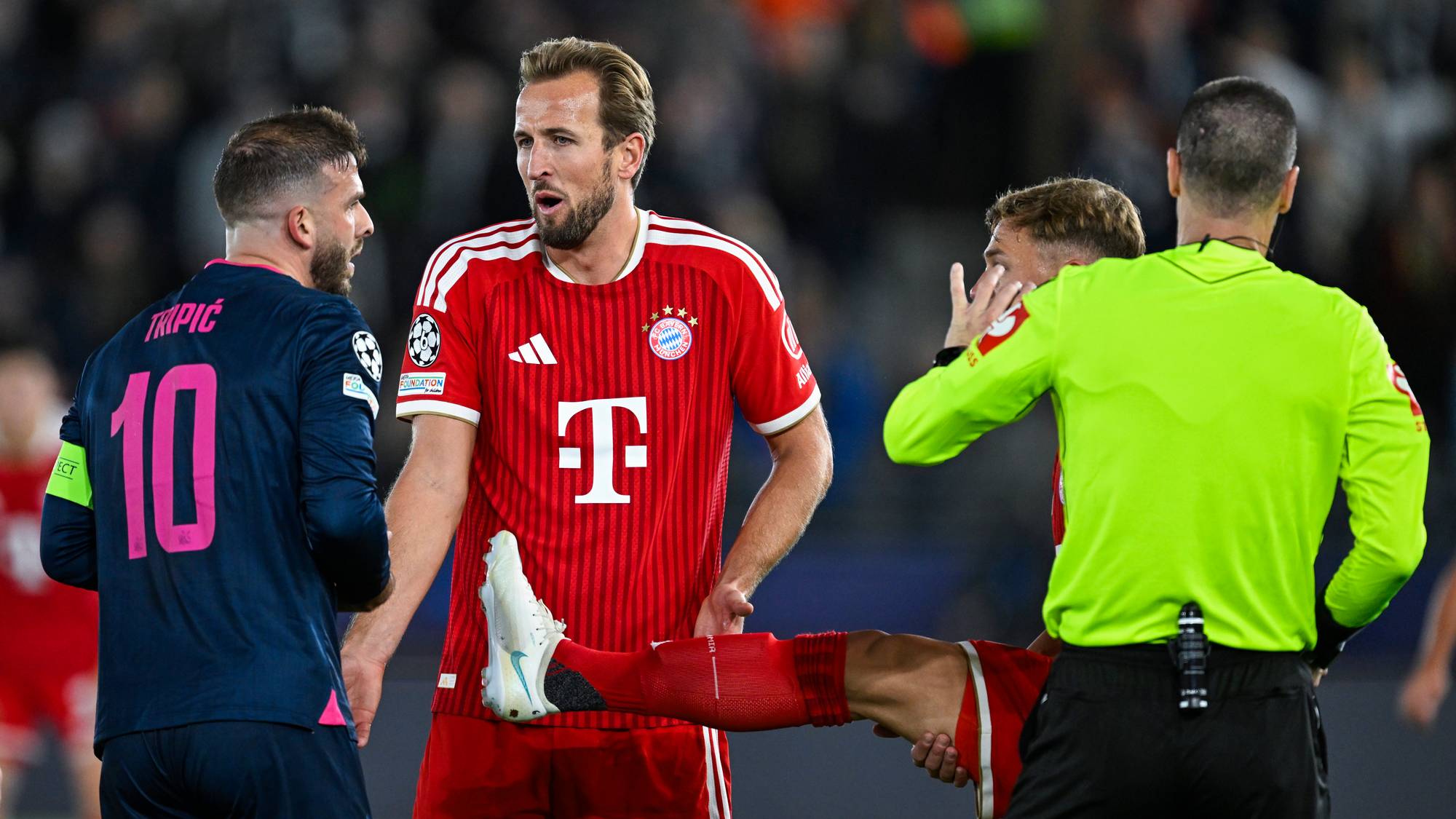 Harry Kane i Bayern München-drakt diskuterer med motspillere og dommer på en fotballbane. (Bildebeskrivelsen er laget av en KI-tjeneste)
