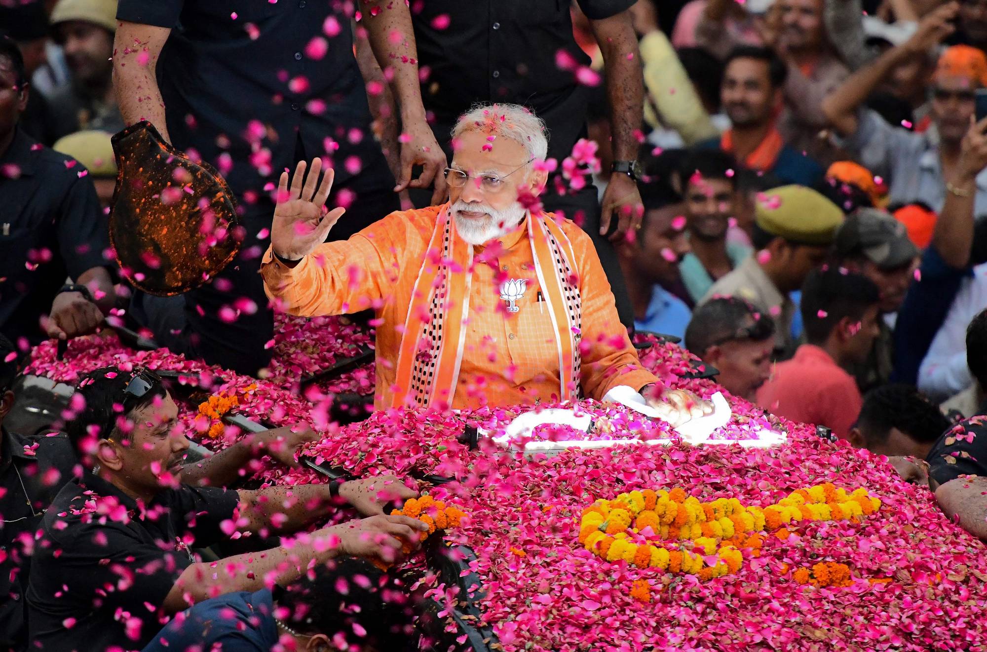 Indias statsminister Narendra Modi vinker til en folkemengde mens han sitter i en bil dekket av blomster. Han er iført en oransje skjorte og sjal. Blomster faller fra luften rundt ham. En stor gruppe mennesker er synlig i bakgrunnen, mange med armene i været. (Bildebeskrivelsen er laget av en KI-tjeneste)
