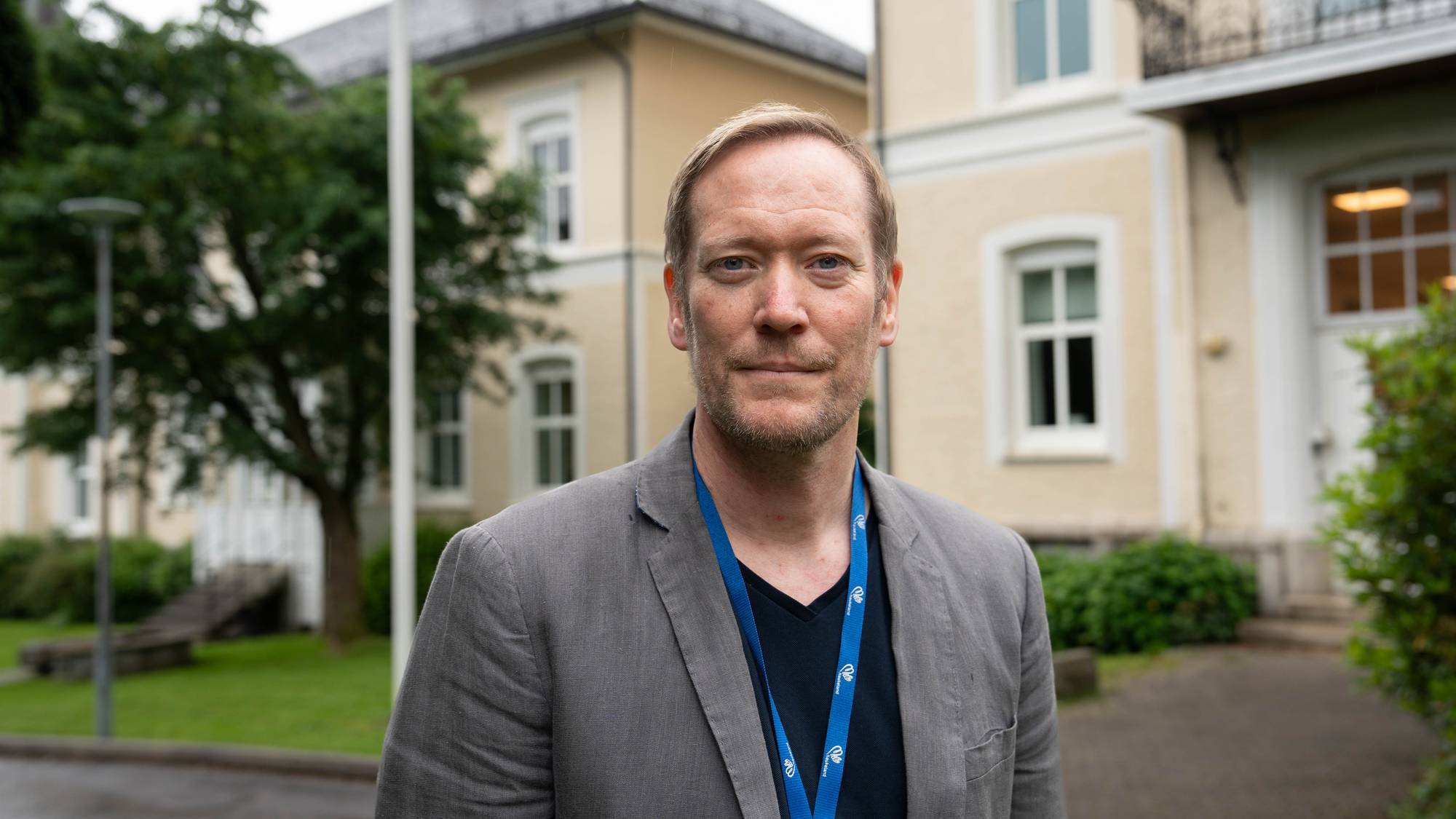 Mann i grå jakke står foran en bygning. Han har et blått nøkkelbånd rundt halsen. 