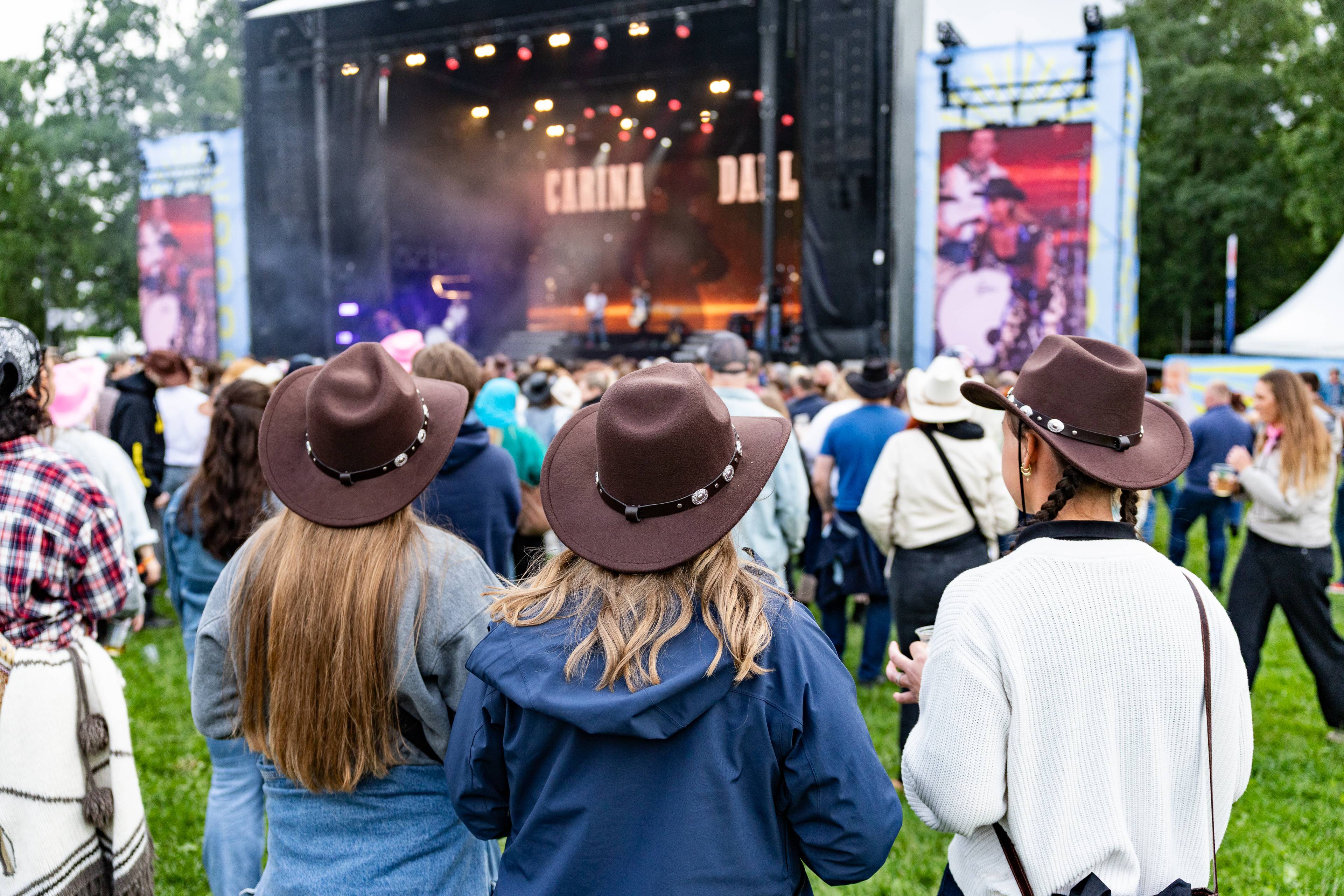 Folkekledte med cowboyhatter står foran en scene på en festival. En gruppe mennesker ser på en konsert med et stort opplyst scenebilde. Flere store skjermer viser også bilder av artister. Festivalområdet er fylt med et stort publikum. (Bildebeskrivelsen er laget av en KI-tjeneste)