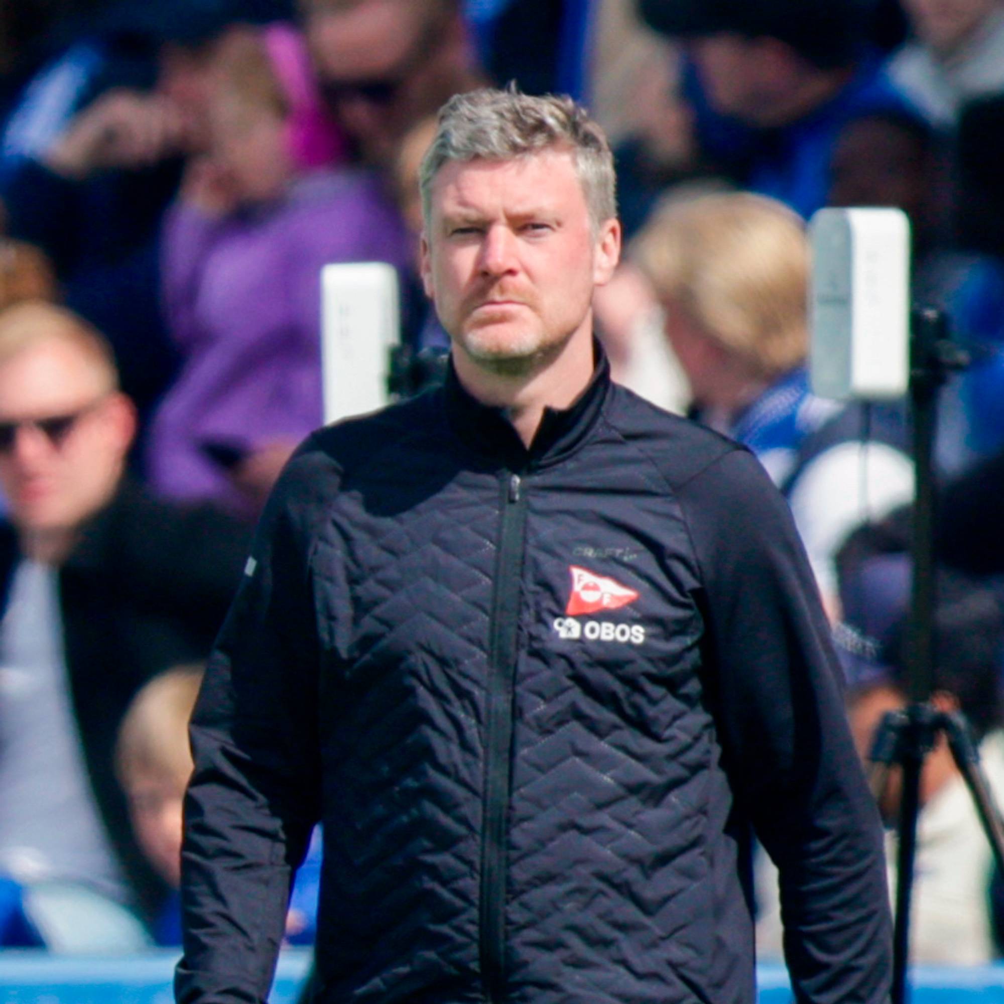 En mann i treningsklær står på et fotballfelt. Bak ham sitter tilskuere på tribunen. En lysreklame med teksten "KIWI mini pris" vises i forgrunnen. (Bildebeskrivelsen er laget av en KI-tjeneste)