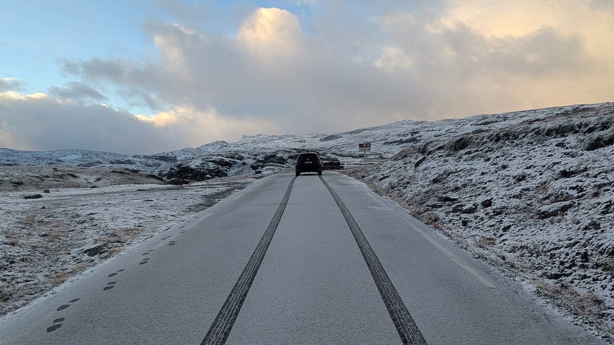 Bil kjører på en vei dekket av et tynt lag snø gjennom et vinterlig fjellandskap. (Bildebeskrivelsen er laget av en KI-tjeneste)