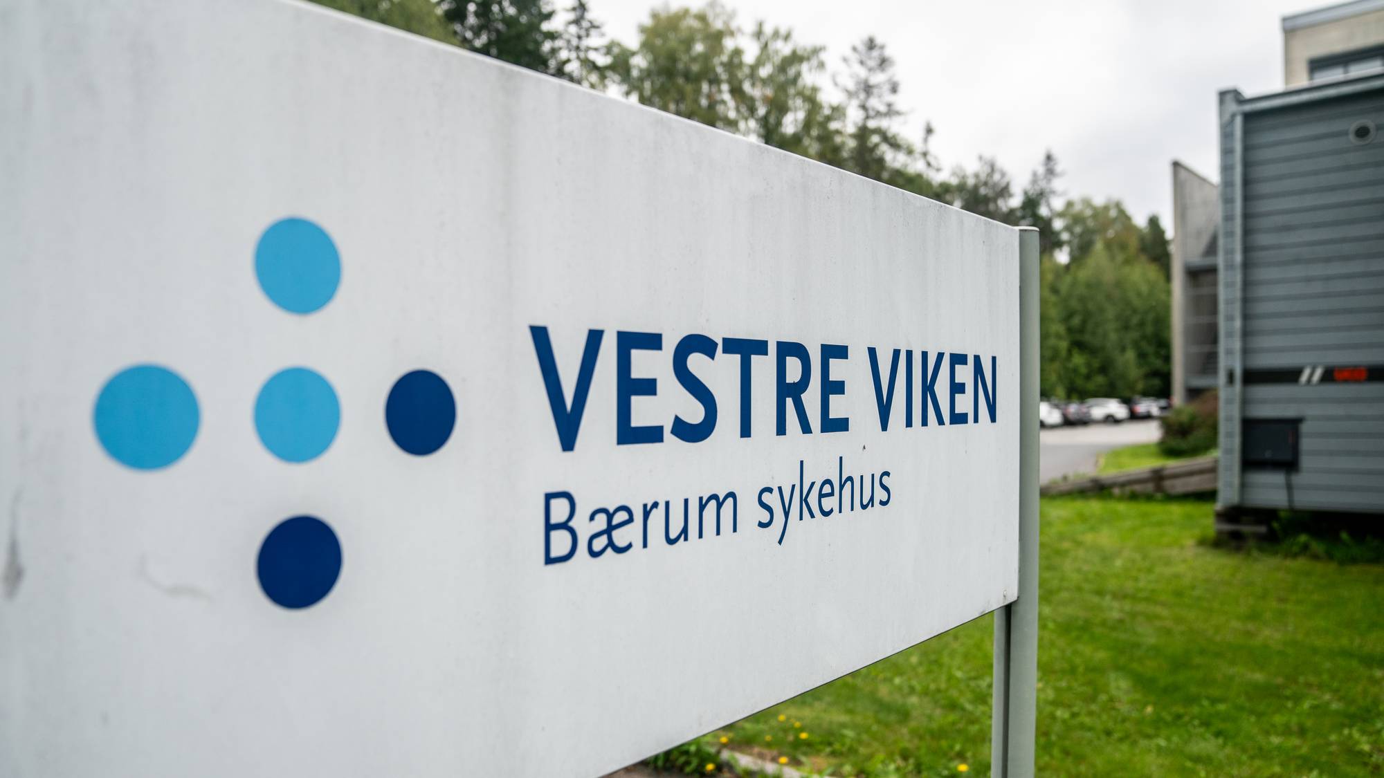 Bilde av et skilt med bygning i bakgrunnen samt trær og gress.