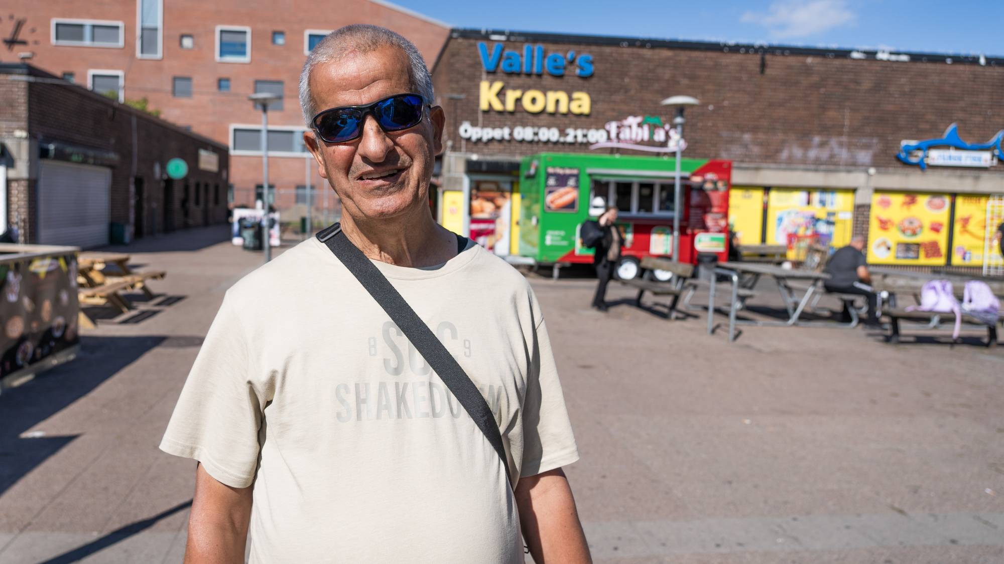 Eldre mann med solbriller står på en plass foran bygget Valles Krona en solfylt dag. (Bildebeskrivelsen er laget av en KI-tjeneste)