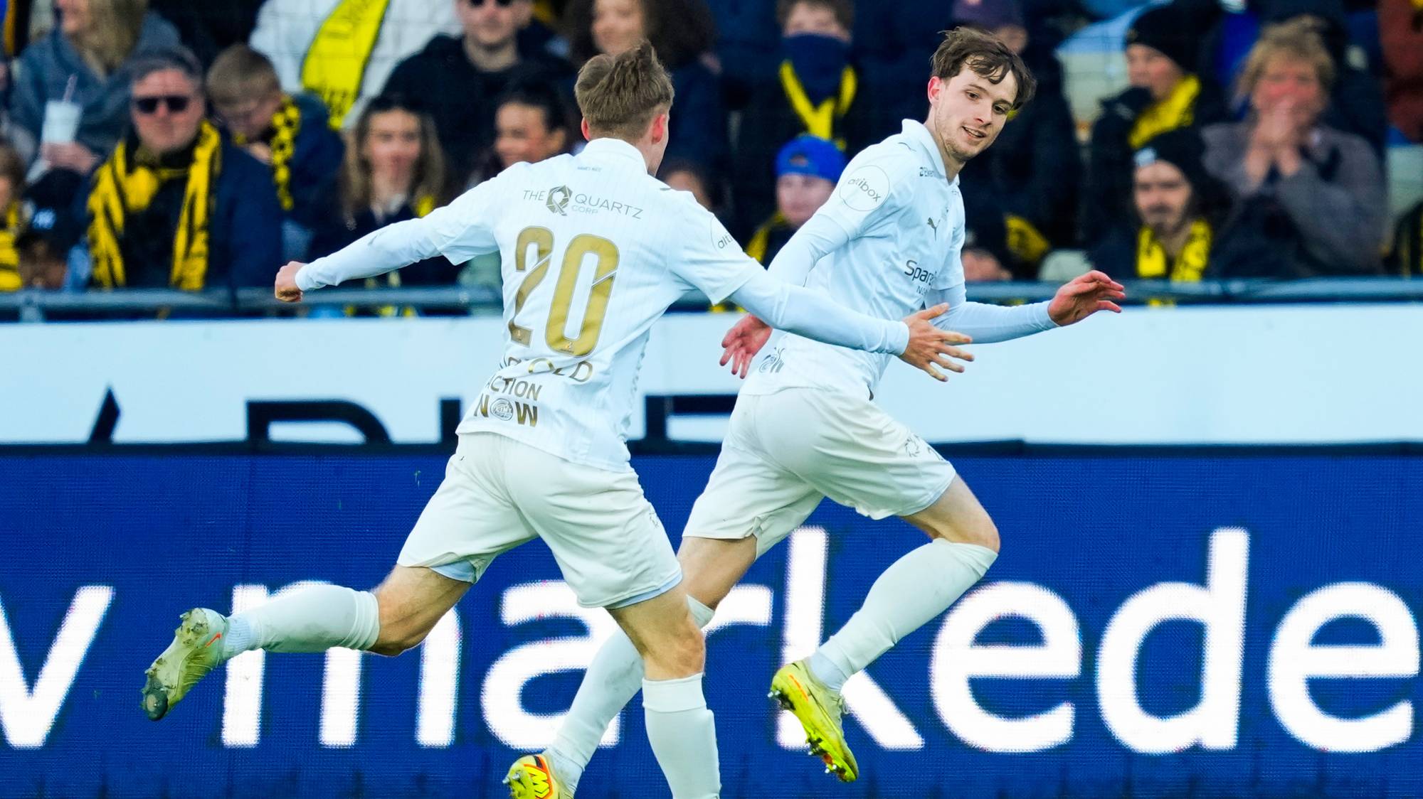 To fotballspillere i hvite drakter løper på en gressbane. En spiller med nummer 20 på ryggen feirer. Tilskuere i gule og sorte drakter sitter på tribunen i bakgrunnen. En stor blå reklameskjerm er synlig bak spillerne. (Bildebeskrivelsen er laget av en KI-tjeneste)