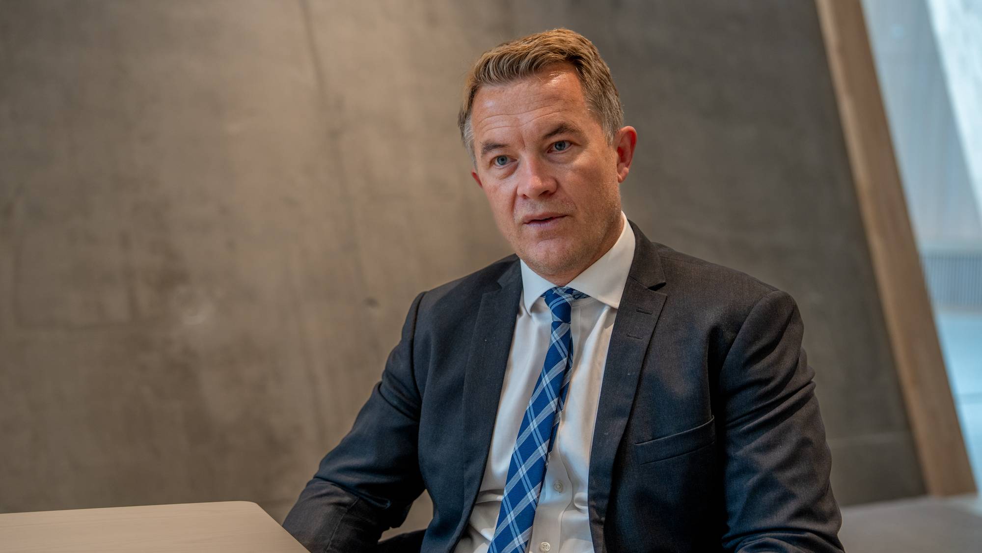 En mann sitter ved et bord foran en betongvegg. Han er kledd i dress og rutete (Bildebeskrivelsen er laget av en KI-tjeneste)