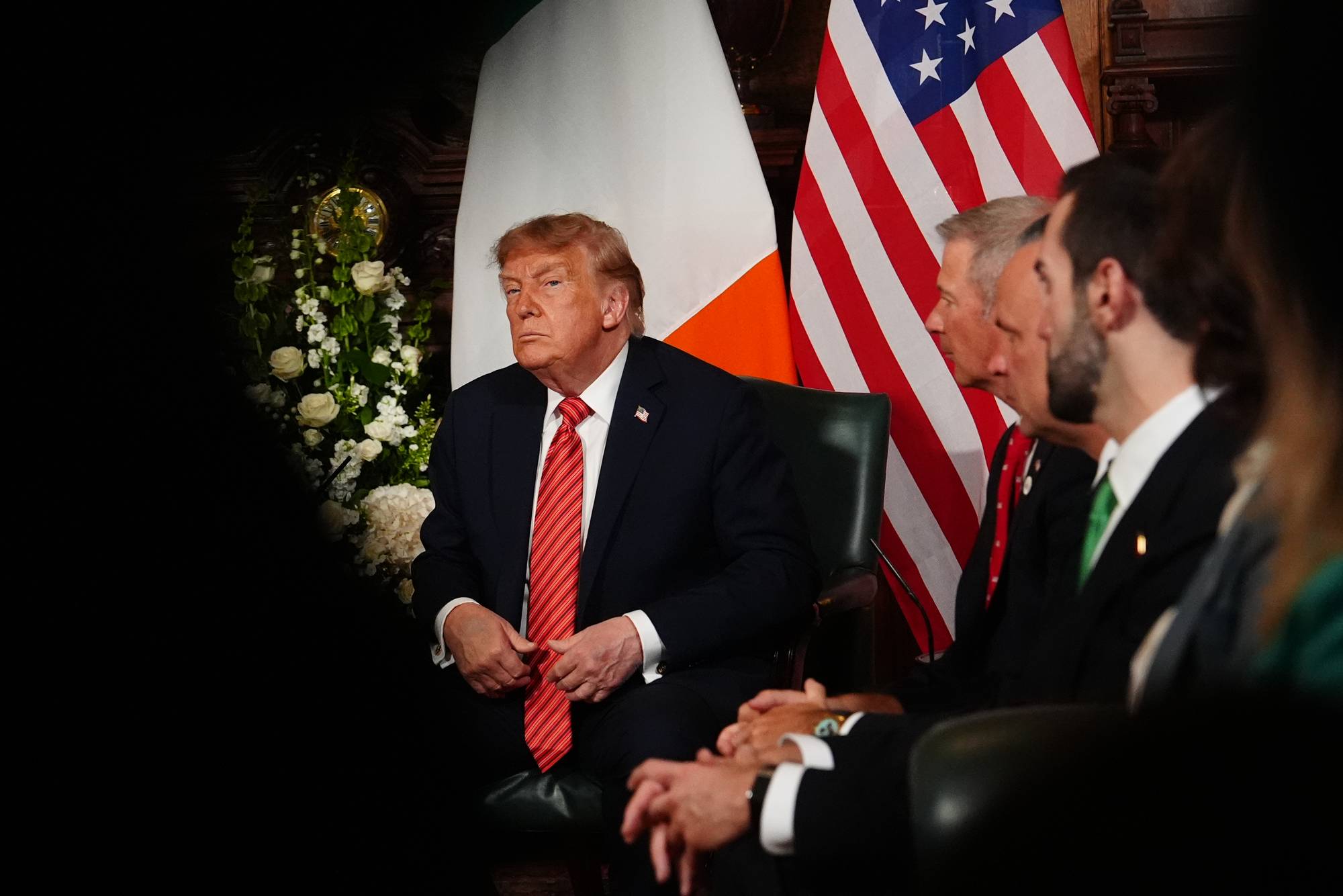 Donald Trump sitter foran irske og amerikanske flagg sammen med andre menn i dress. (Bildebeskrivelsen er laget av en KI-tjeneste)