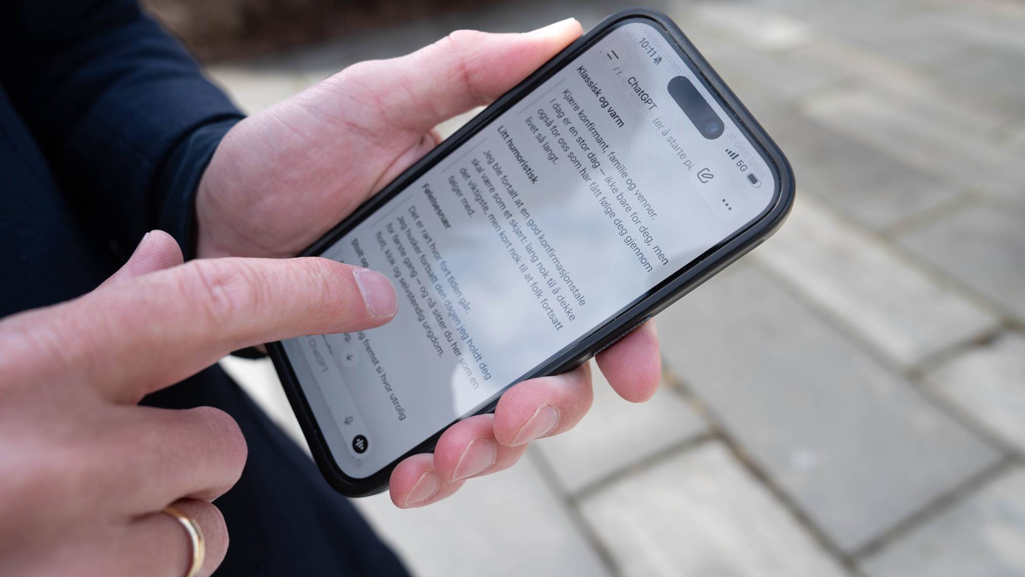 Eit nærbilete av handa til taleekspert Trond Albert Skjelbred. Han held ein mobiltelefon, og peikar med peikefingeren på skjermen. Mobiltelefonen står open på appen Open AI, som viser eit KI-generert forslag til ein konfirmasjonstale. 