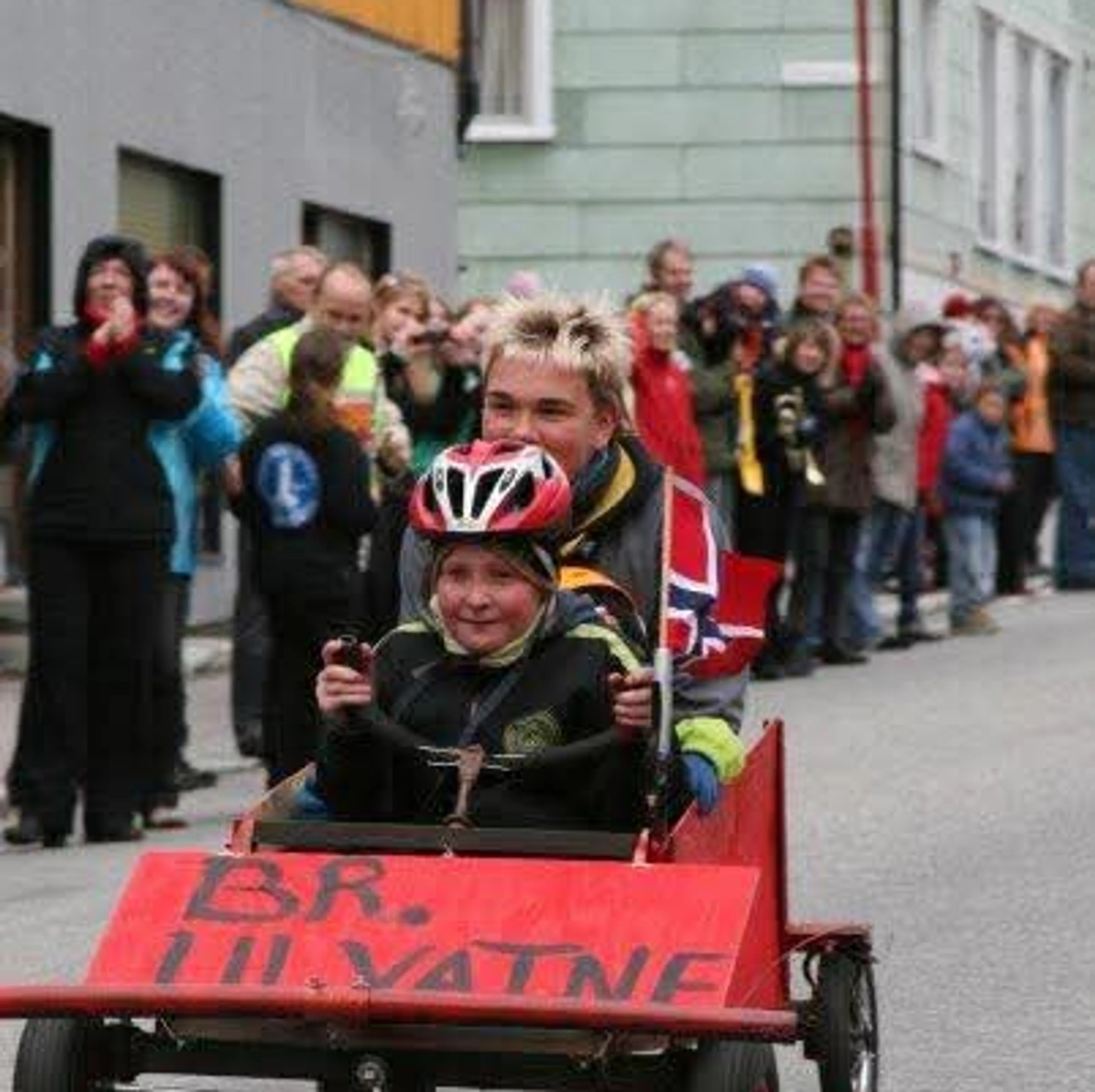 To brødre sitter i en ola-bil som er malt rød og merket Br. Ulvatne. Kjører nedover bakke i Honningsvåg med flagget vaiende på siden.