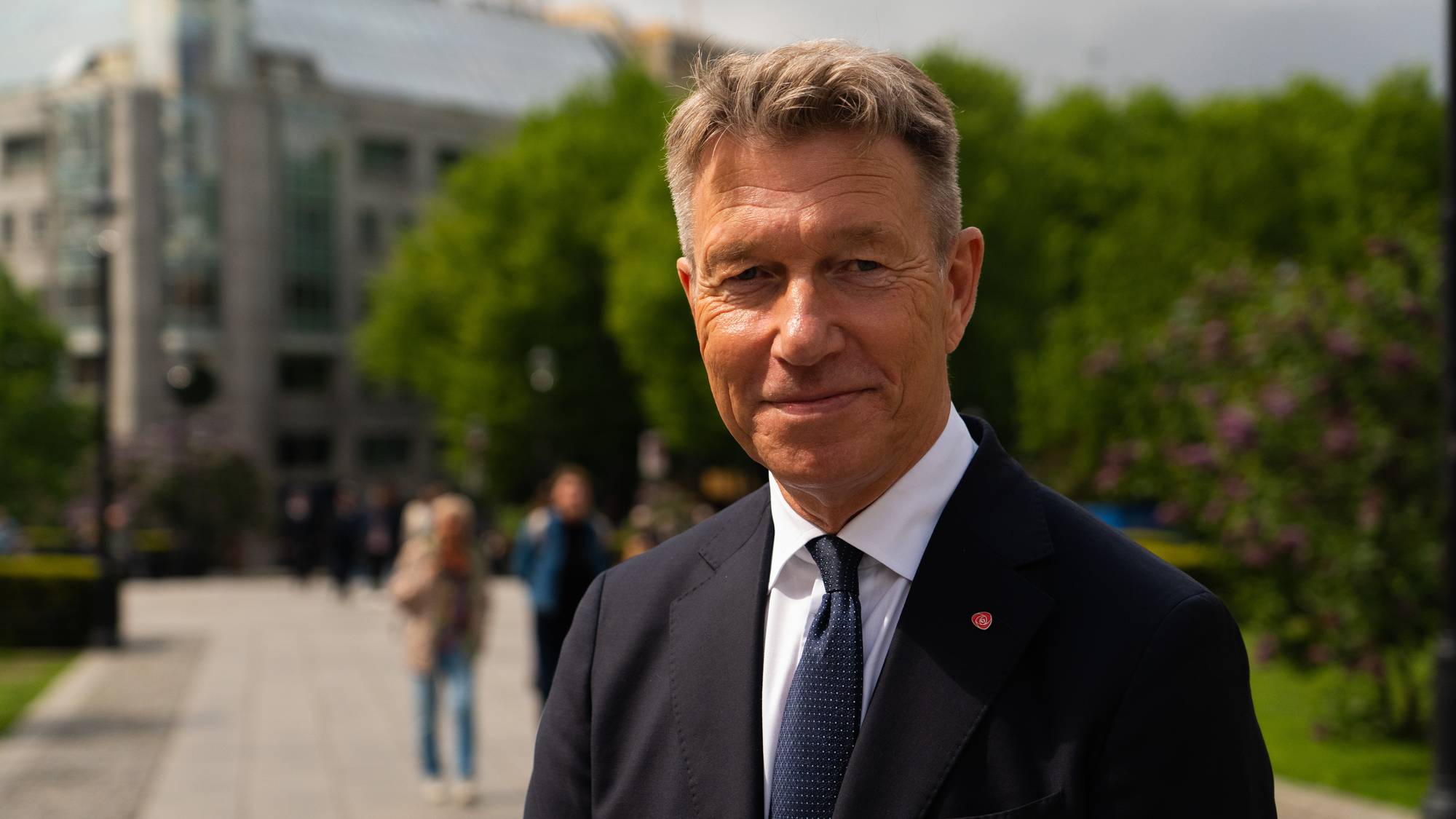 Portrett av Terje Aasland, energiminister. Står utendørs i bybildet. Trær og mennesker er uskarpe i bakgrunnen. Det er sol og han smiler. 