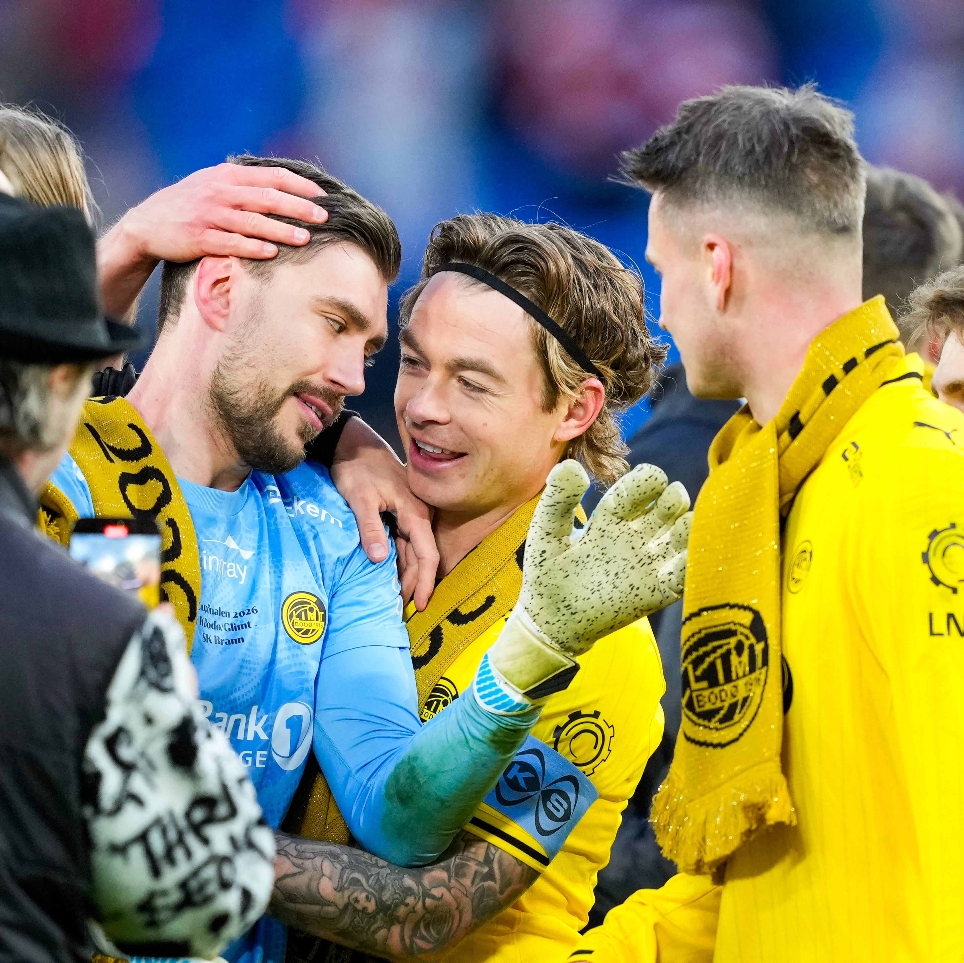 Fotballspillere i gule og lyseblå drakter feirer på en bane. En spiller med keeperhanske og supporterhatt tar bilde av spillerne med mobil. Mange supportere i bakgrunnen. (Bildebeskrivelsen er laget av en KI-tjeneste)