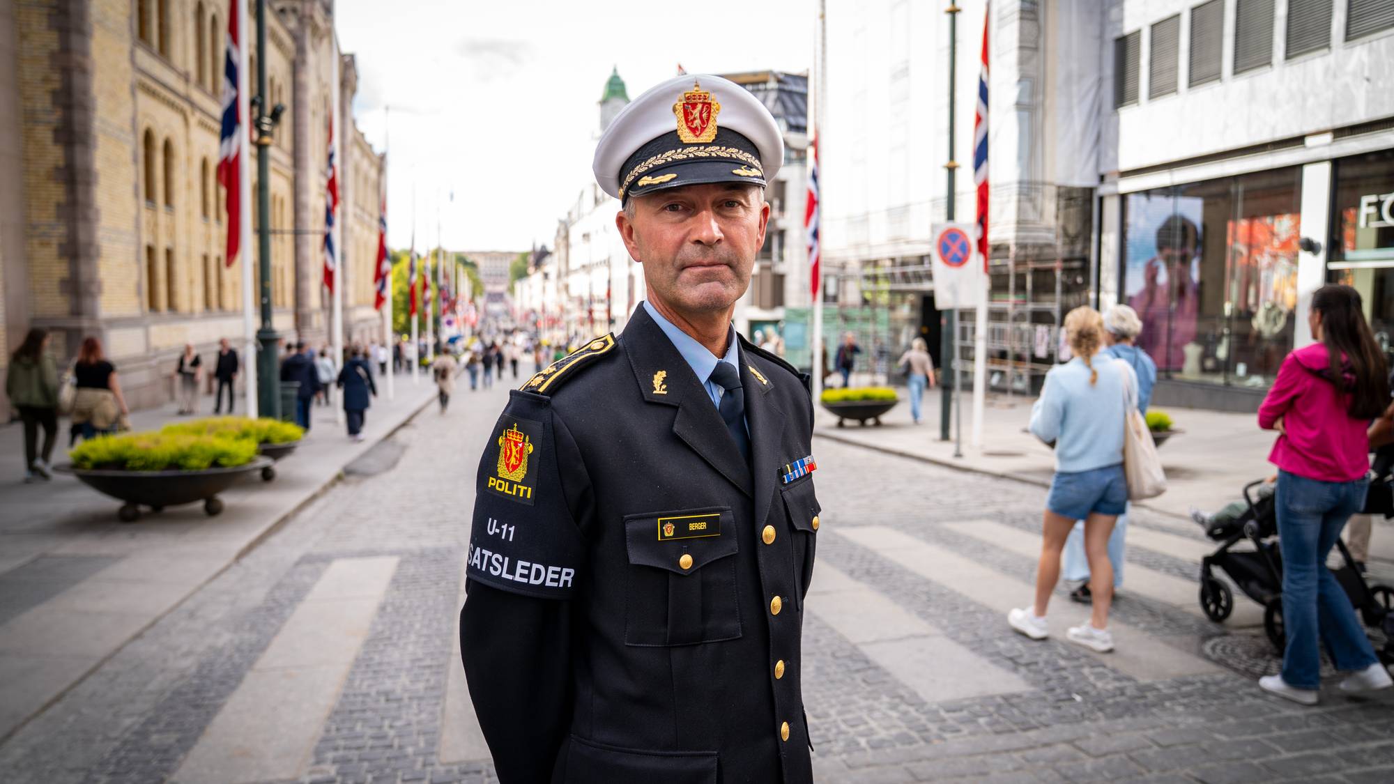 Innsatsleder fra politiet i uniform står i en bygate med flagg og fotgjengere i bakgrunnen. (Bildebeskrivelsen er laget av en KI-tjeneste)