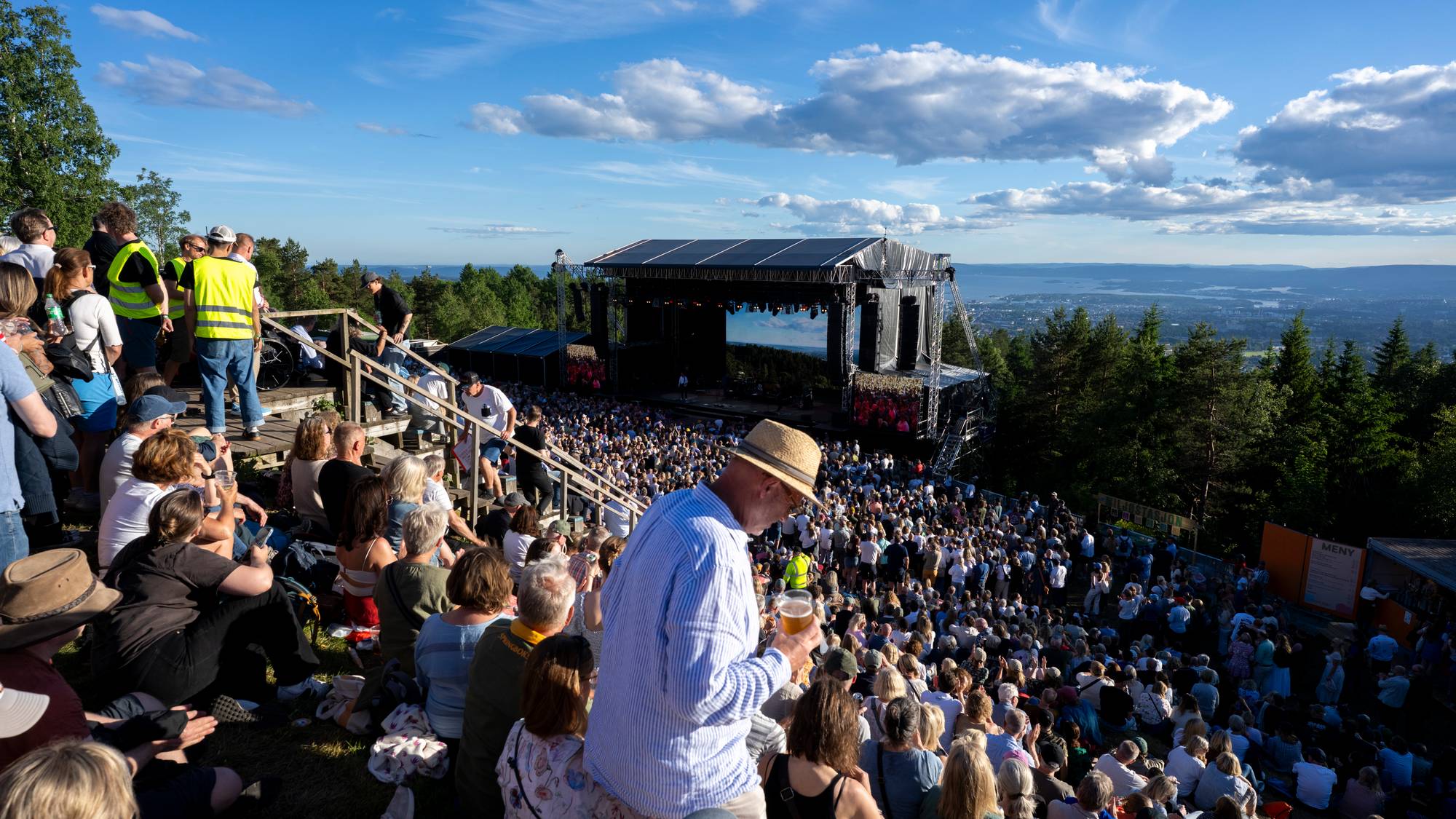 En stor folkemengde ser på en konsert på et utendørsarrangement. En scene med lyd- og lysutstyr er sentralt plassert. Mange tilskuere sitter på tribuner eller på gresset. (Bildebeskrivelsen er laget av en KI-tjeneste)
